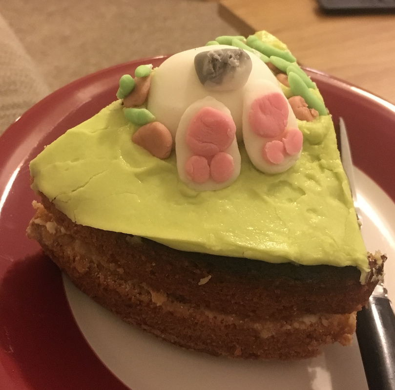 Slice of a decorated cake I've baked. It's a standard victoria two tier sandwich cake. The cake is decorated with a green butter icing base. On the cake is the rear end of a rabbit sticking out of mud made out of fondant. The cake is sat on a red plate with a table and carpet in the background