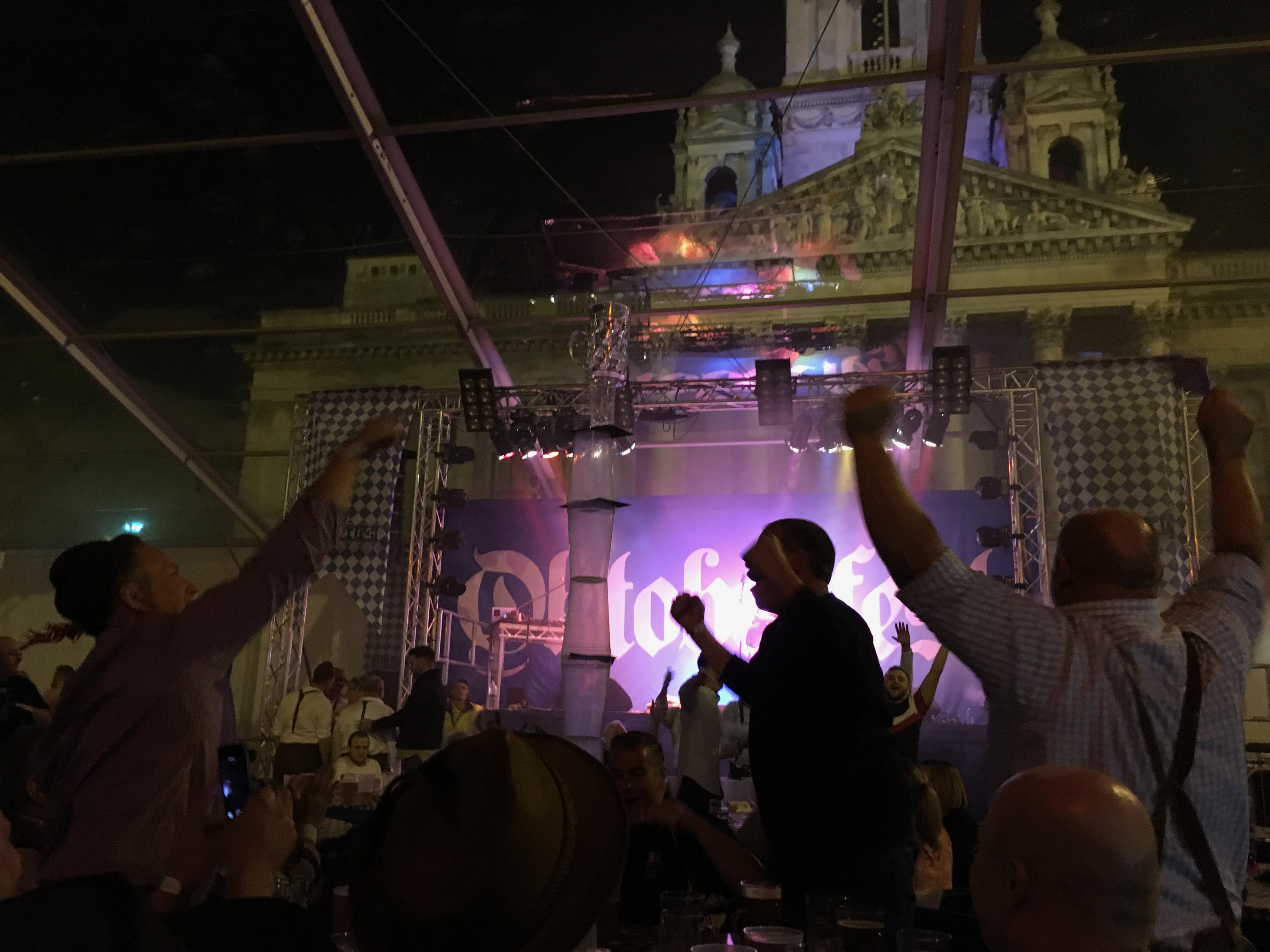 A table with many glasses stacked higher than the people standing around, likely over 7 feet tall. The person on the left is pointing to it and the people on the right is celebrating with their arms raised. In the background is the stage with lighting and a blue and white Oktoberfest background