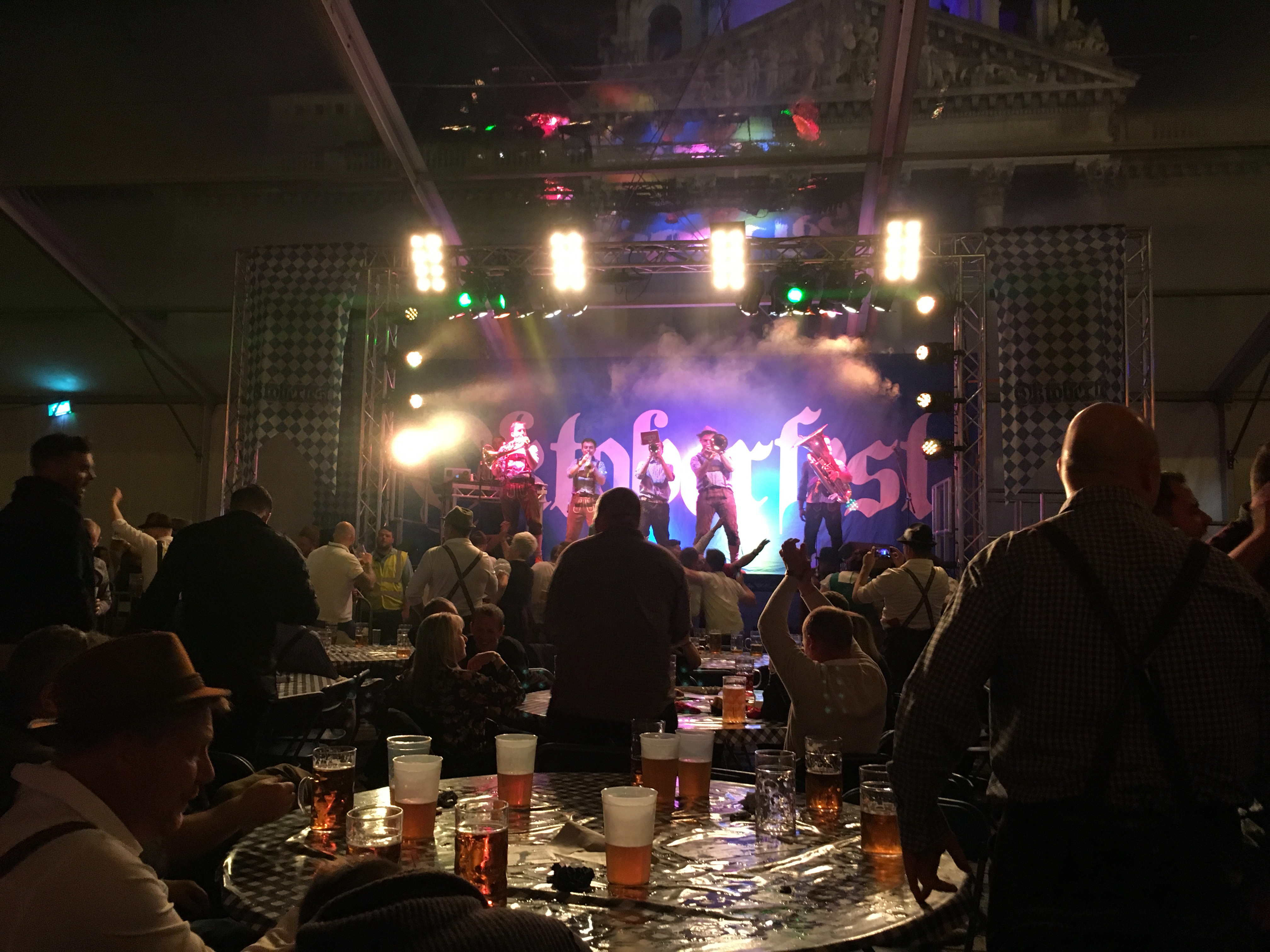 Oompah band on stage with blue and white background spelling out Oktoberfest. The stage is surrounded by lights. In front of the stage is several tables filled with various glasses of beer. Several people are stood up singing and dancing.