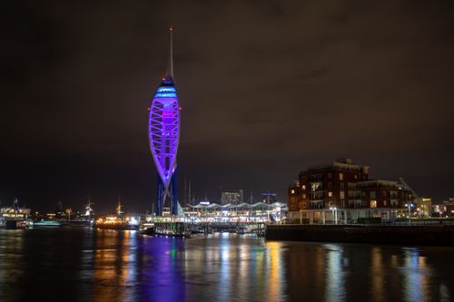 Spinnaker Tower viewed from behind in Old Portsmouth at hightide tide at night. The long exposure has caused the sea to be smooth. The tower is lit up purple.