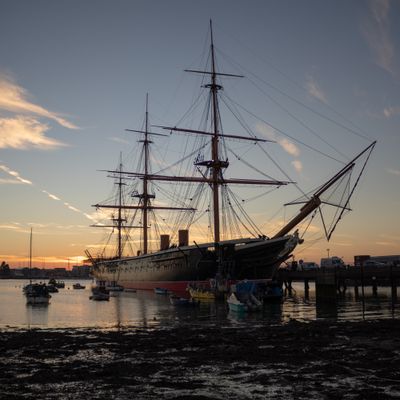 HMS Warrior in Portsmouth Harbour at low tide with the setting sun behind. Beach and seaweed in the foreground.