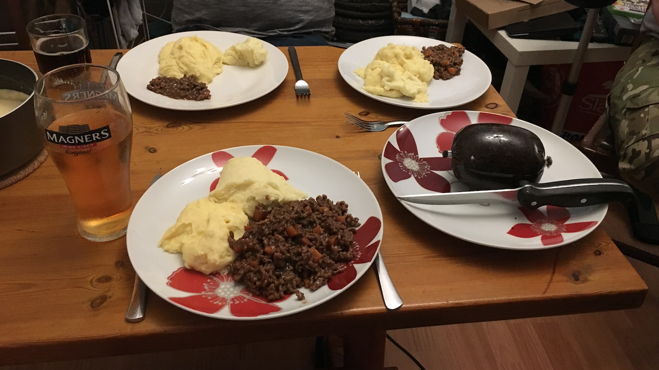Table with 4 plates on them. 3 of the plates have mash and some stew made of beef and various vegetables. On the other plate is a small sealed haggis and a steak knife