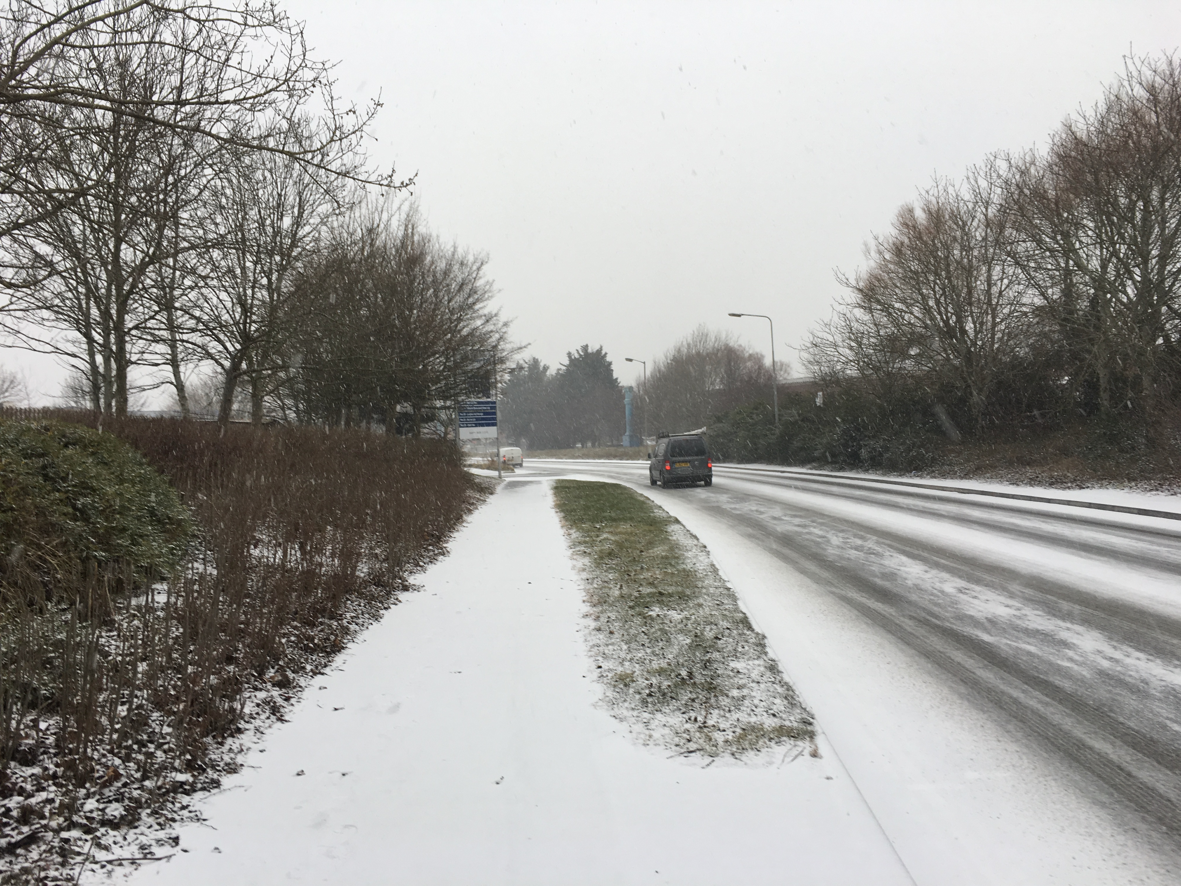 There's a pathway on the left that is completely covered in snow. On the right is a road which has snow on it and lines where cars have been driving. There is visible flakes of snow in the air