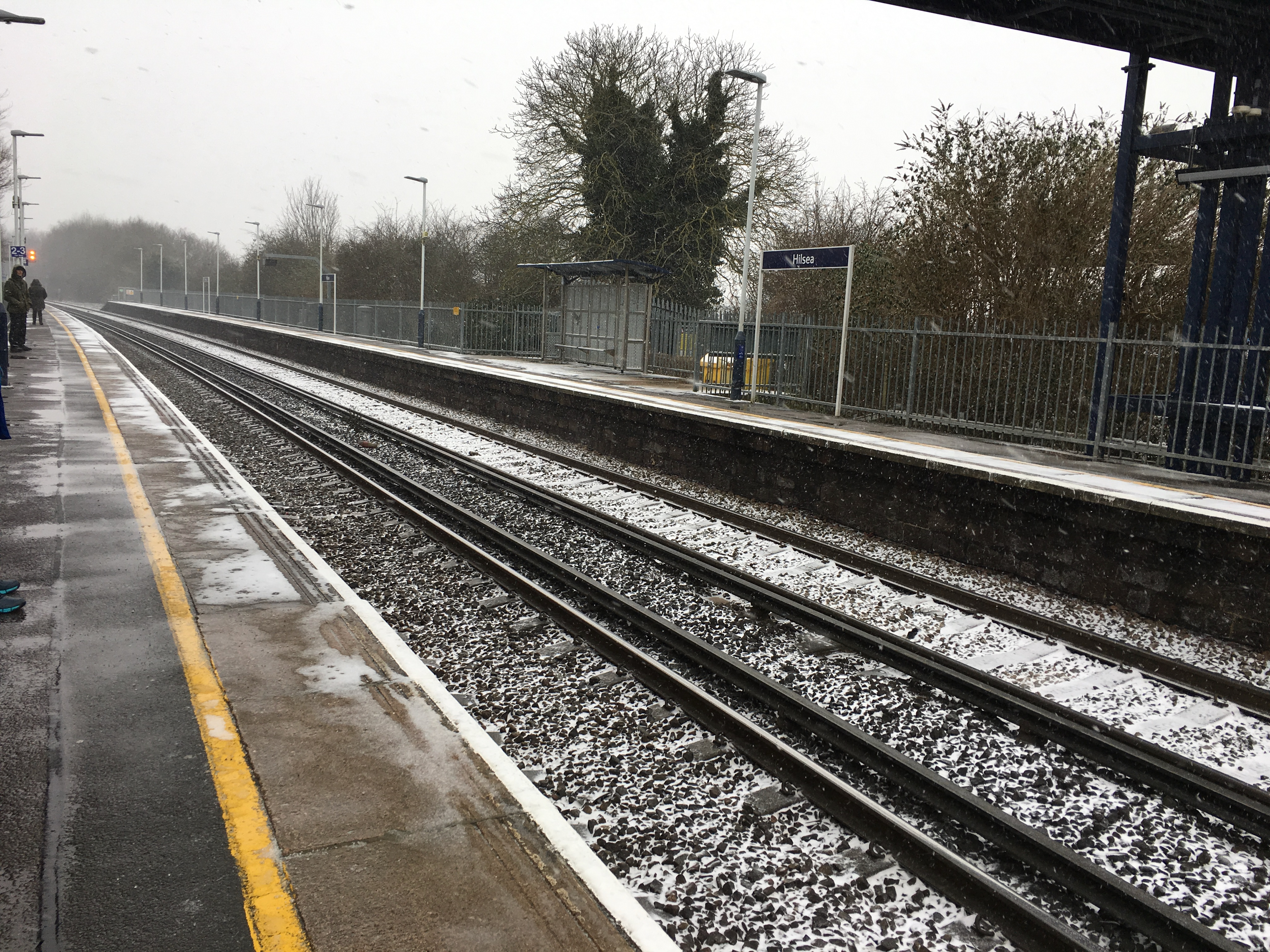Railway station with two tracks and a platform either side. On the platforms and track ballast there is snow. There is snow flakes visible in the air going diagonally