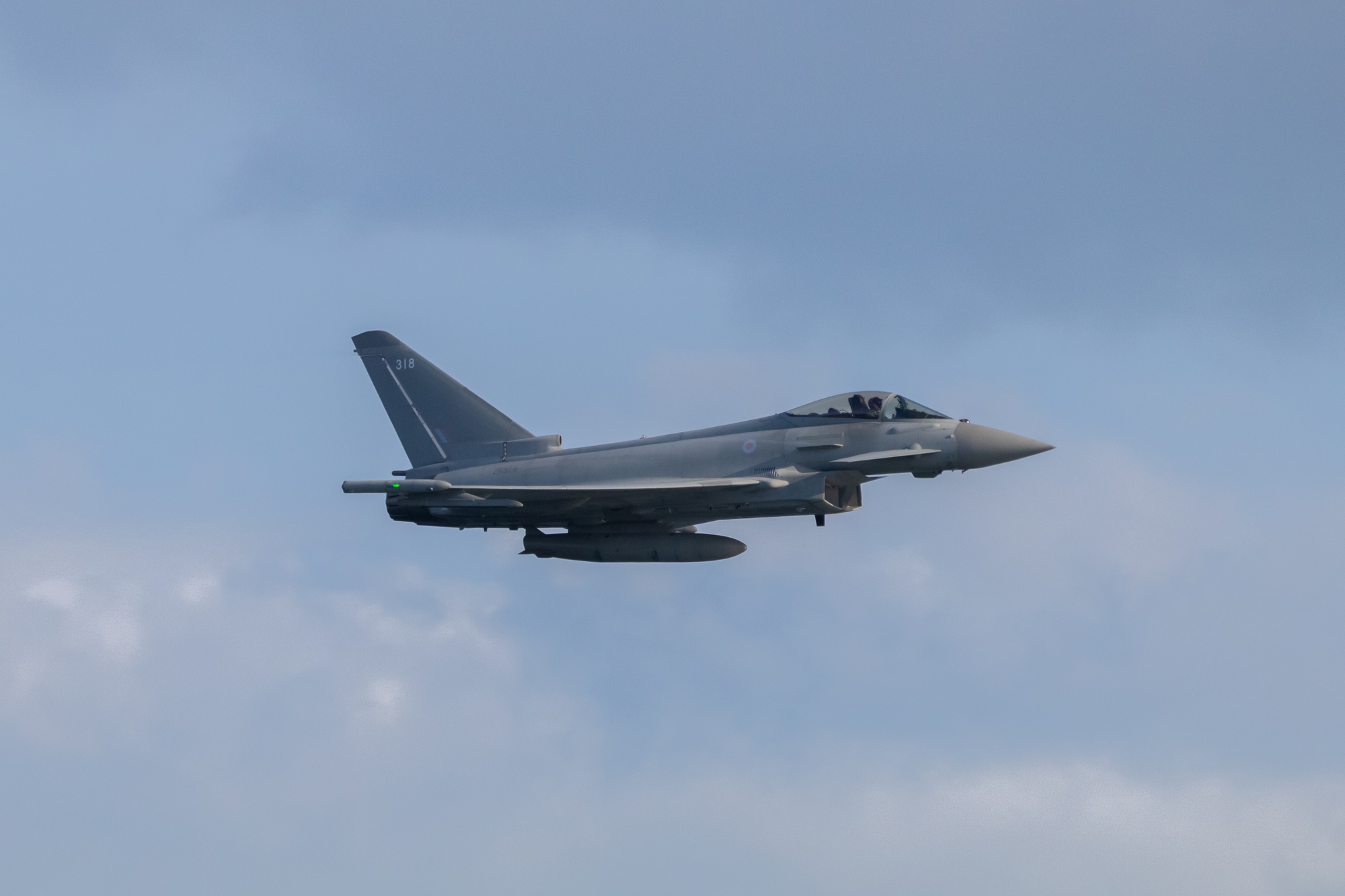 Eurofighter Typhoon flying to the right of frame against blue sky. The aircraft is in standard grey livery with a fake warhead on the underside of the fuselage