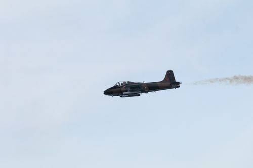 Strikemaster flying to the left of frame against grey sky. The eginle exhausts a noticable brown colour. The plane is in dark green and brown livery