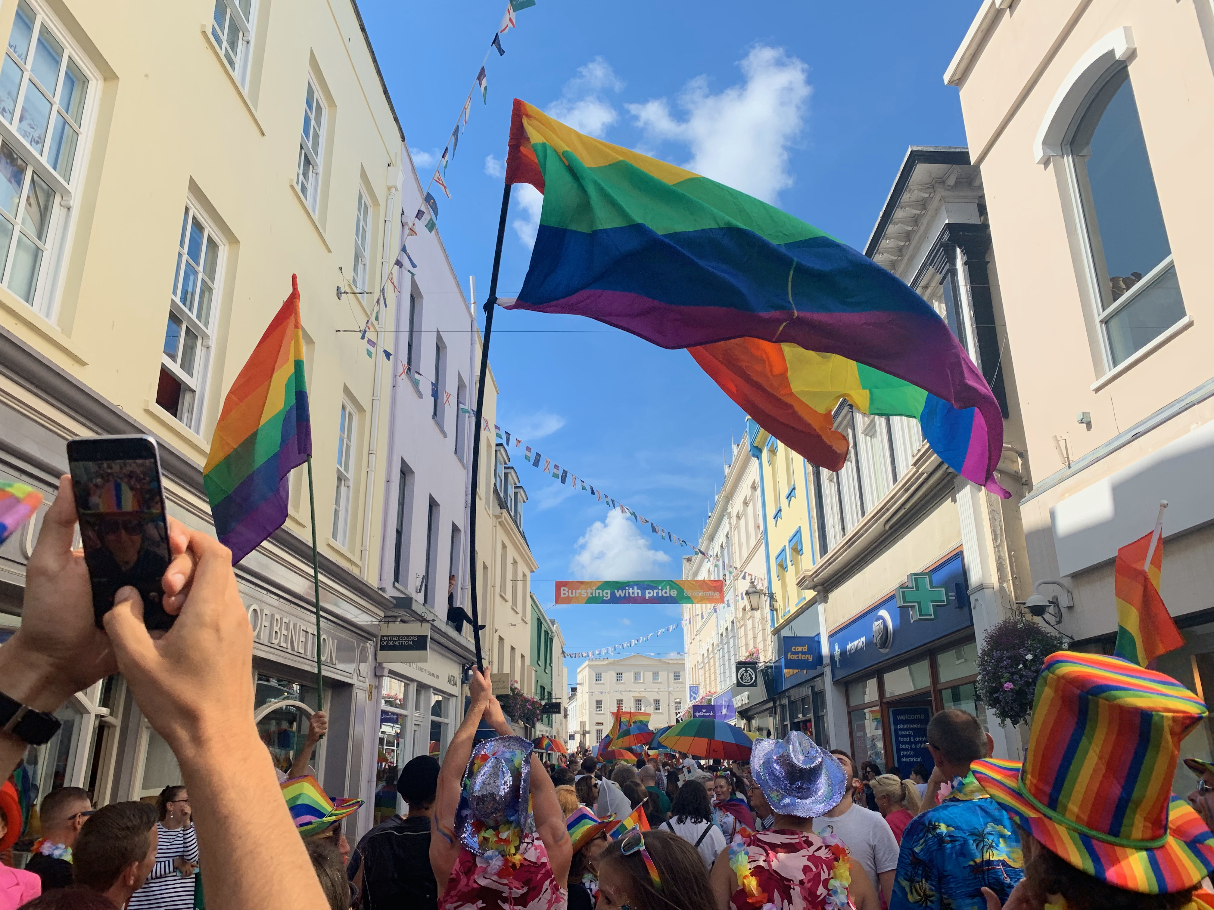 Jersey Price march through the high street. Many people in a crowd wearing brightly coloured clothing. Several people are waving pride flags and holding pride umbrellas
