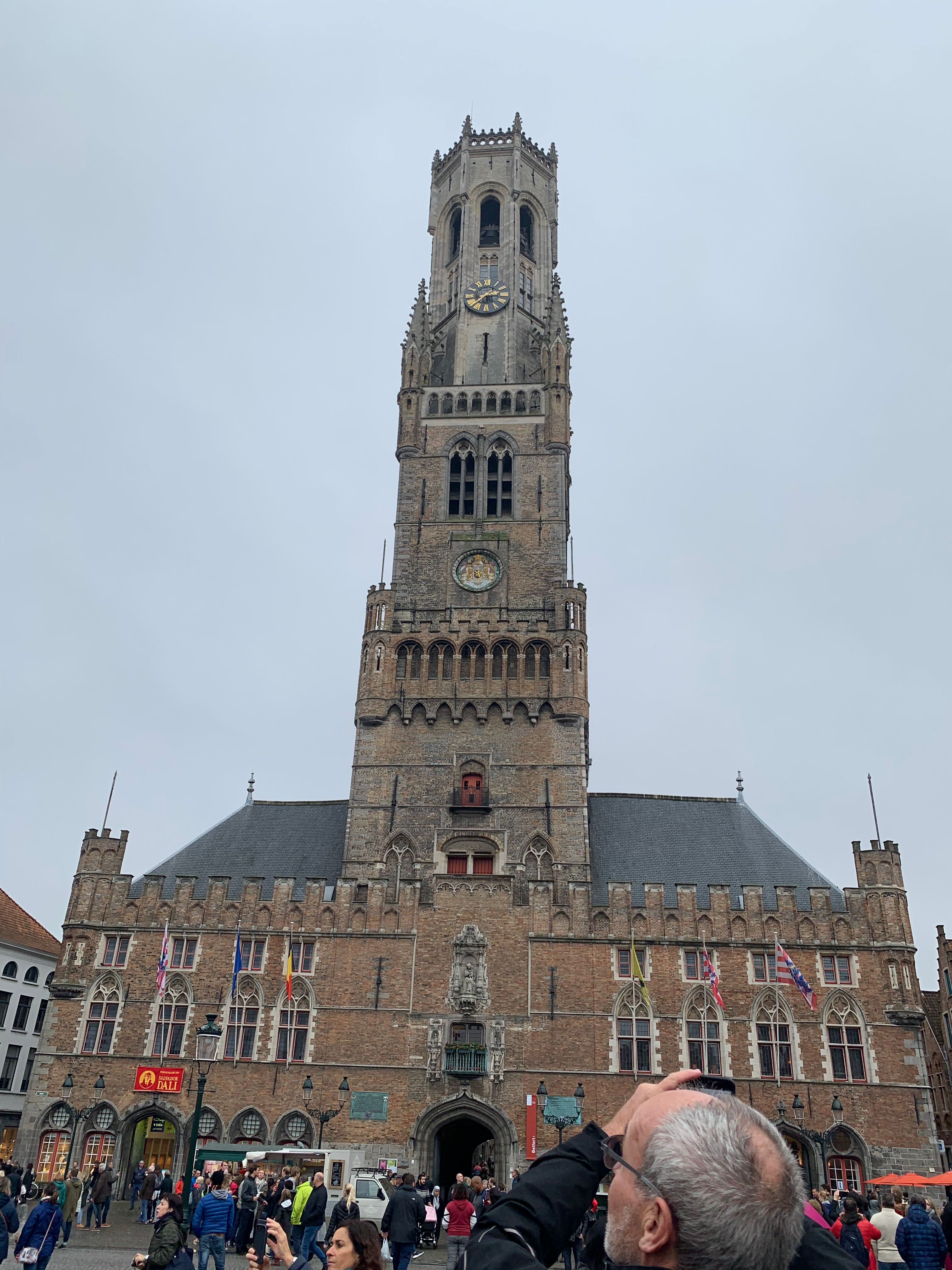 Large and tall clock tower in the Grote Markt in Bruges. It is a tall dark brown/grey brick tower. The top part is octagonal in design with a small clock. The lower parts of the tower is of square design and has a crest. The lower part of the building is a more red/brown brick. There are many people in the square in front of the tower