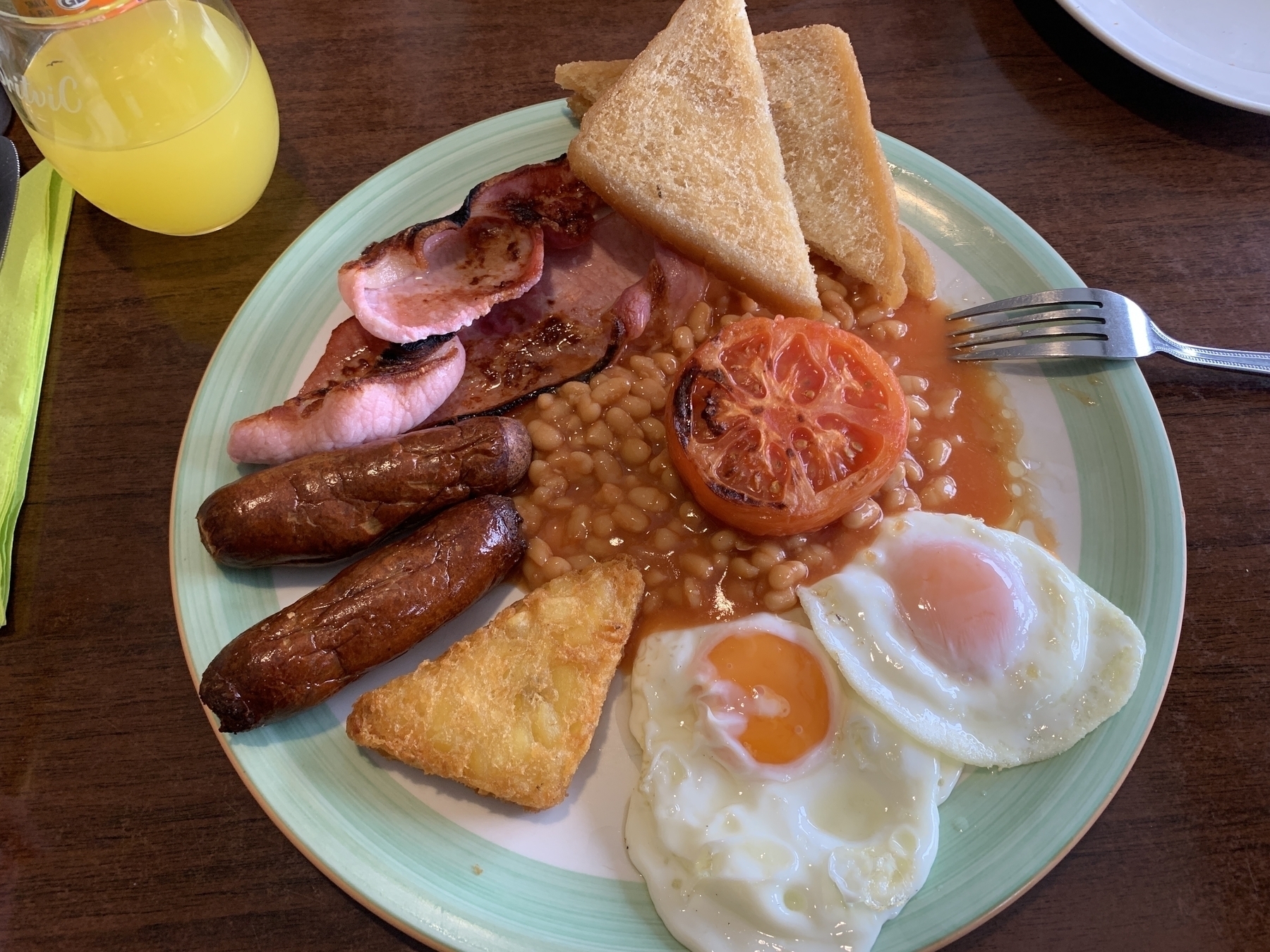 Large traditional breakfast on a blue/green plate. The breakfast includes 2 slices of fried-bread, 2 sunny-side up eggs, 1 hash-brown, 2 sausages, 2 slices of bacon, half a large fried tomato and a large portion of baked beans. My fork is sitting on the right side of the plate. In the background is a glass of fanta and a dark wooden table