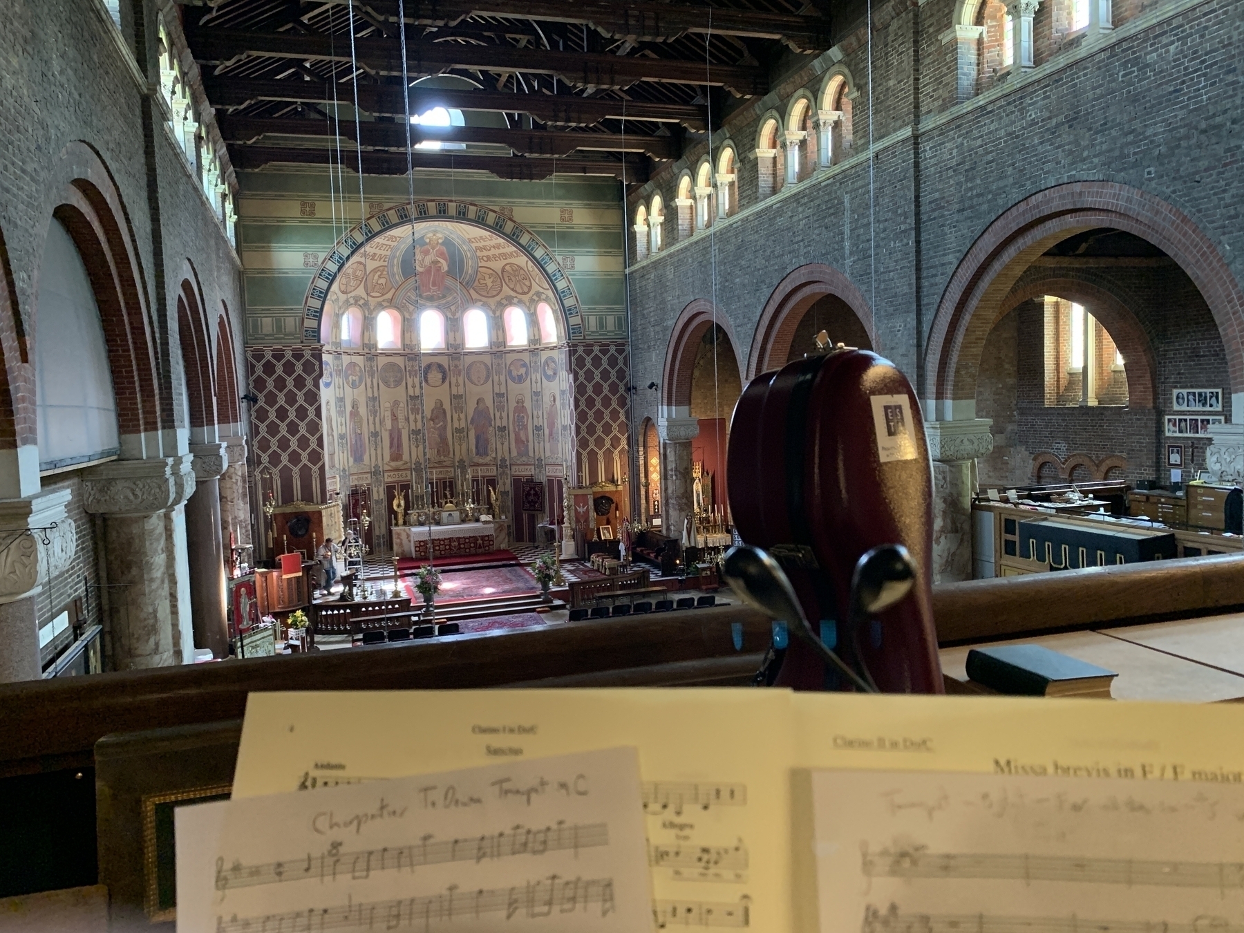 View from a balcony into a large church. The church has very high ceilings with the altar in an enclosed semi-circle at the end of the hall. In the foreground is some music and a cello case