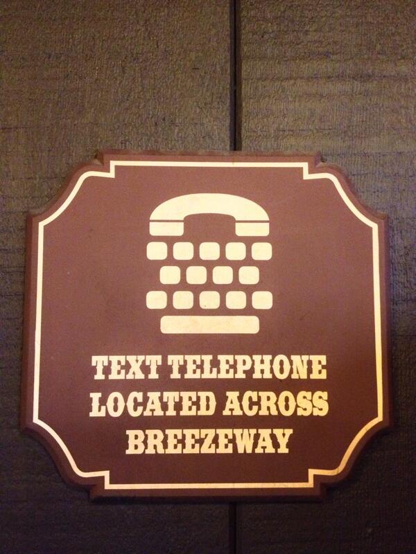 Brown sign reading "Text telephone located across breezeway". Above the text is a phone icon with several buttons below it arranged roughtly like a keyboard and space bar
