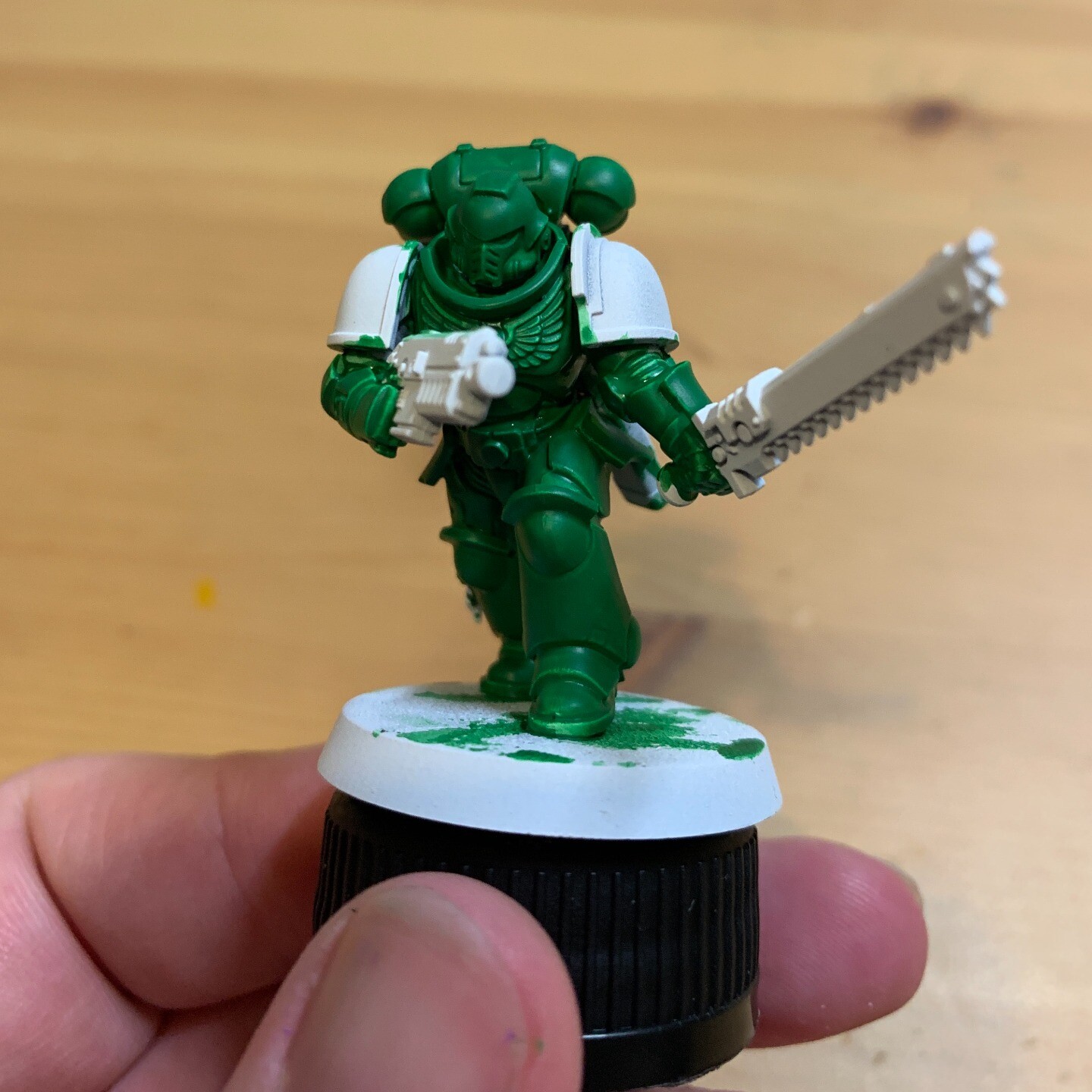 I'm holding a half-painted Warhammer 40k Assult Intercessor Space Marine. The main armour and backpack have been painted in a middle-to-dark green colour. The shoulder pads and weapons are unpainted and white. The base is unpainted and is on a black bottle cap.  My hand is in the bottom of the frame with some green paint on the nail. The background is white parchment.