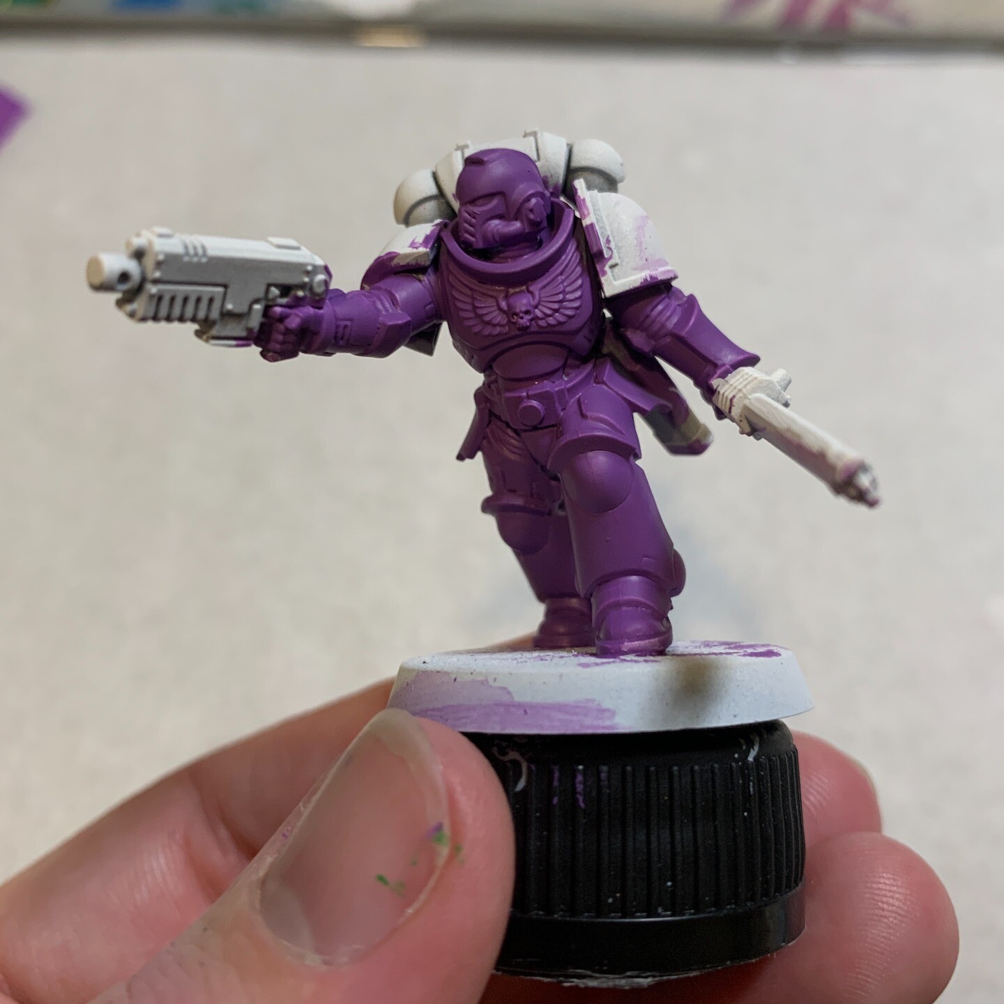 I'm holding a half-painted Warhammer 40k Assult Intercessor Space Marine. The main armour has been painted in a pink-purple colour. The backpack, shoulder pads and weapons are unpainted and white. The base is unpainted and is on a black bottle cap.  My hand is in the bottom of the frame with some purple and green paint on the nail. The background is white parchment.