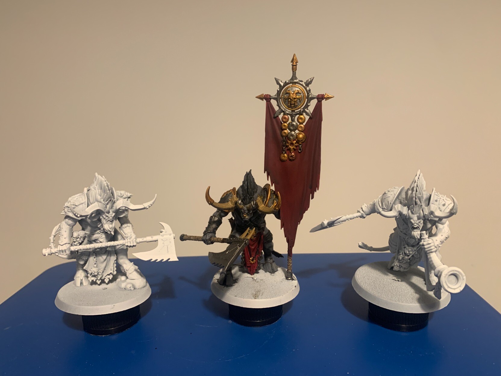 Three Ogroid Theridons. The two outside ones are unpainted and primed in white. The middle one is painted in dark greys with gold trim and is holding a dark red banner. The banner is topped by both silver and gold metal elements.