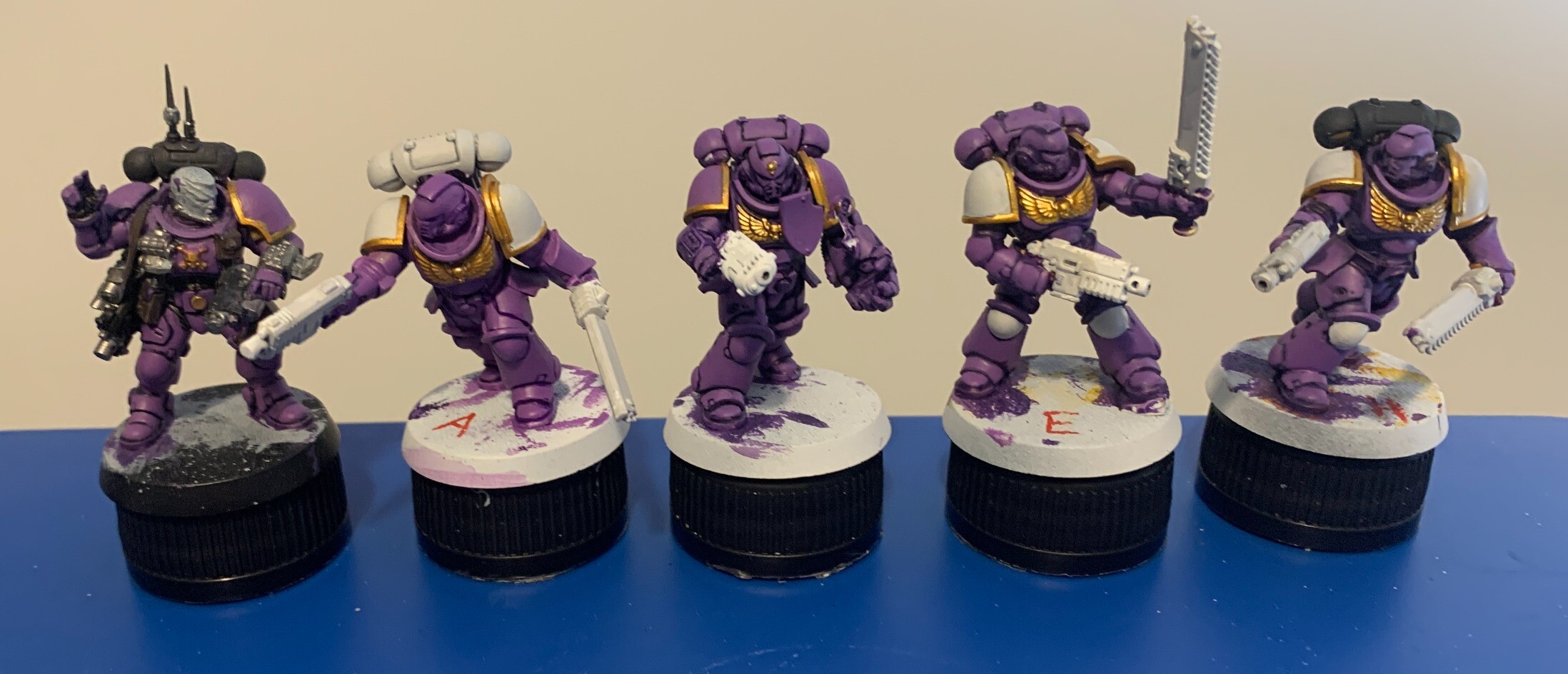 5 space purple space marines in a line with variations of accent colours. The purple is a middle brightness and appears warm, leaning more red than blue. The trim of the armour is gold. From left to right the variations are: purple pauldrons and black backpack. White pauldrons and white backpack. Purple pauldrons and purple backpack. White pauldrons and knee pads, and a purple backpack. White pauldrons and knee pads with a black backpack. The models are sat on black bottle caps on top of a blue plastic lid.