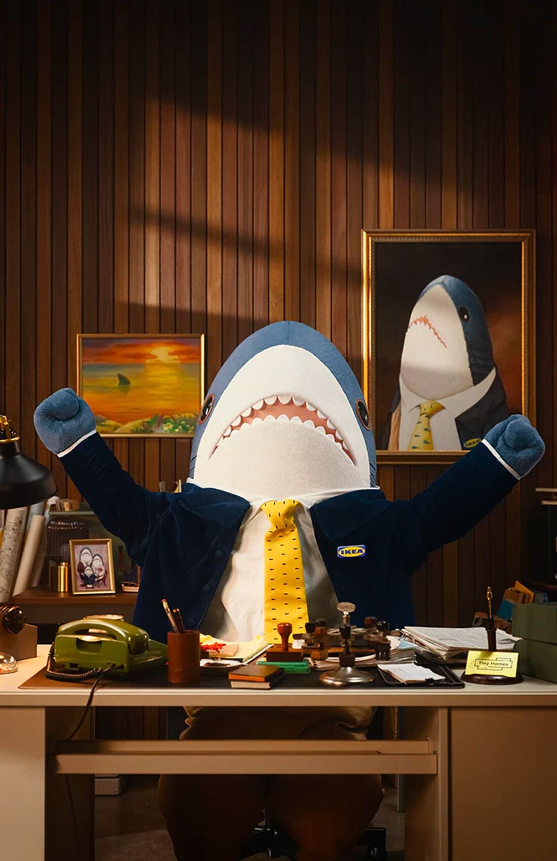 A full-size Blahaj (shark) mascot suit sitting at a desk. The shark is sitting down at the desk with its arms raised in triumph. The Blahaj is wearing light brown trousers, a dark blue jacket and a yellow tie. The desk is littered with various stationary items. The wall behind is vertical wood panelling with various paintings of sharks