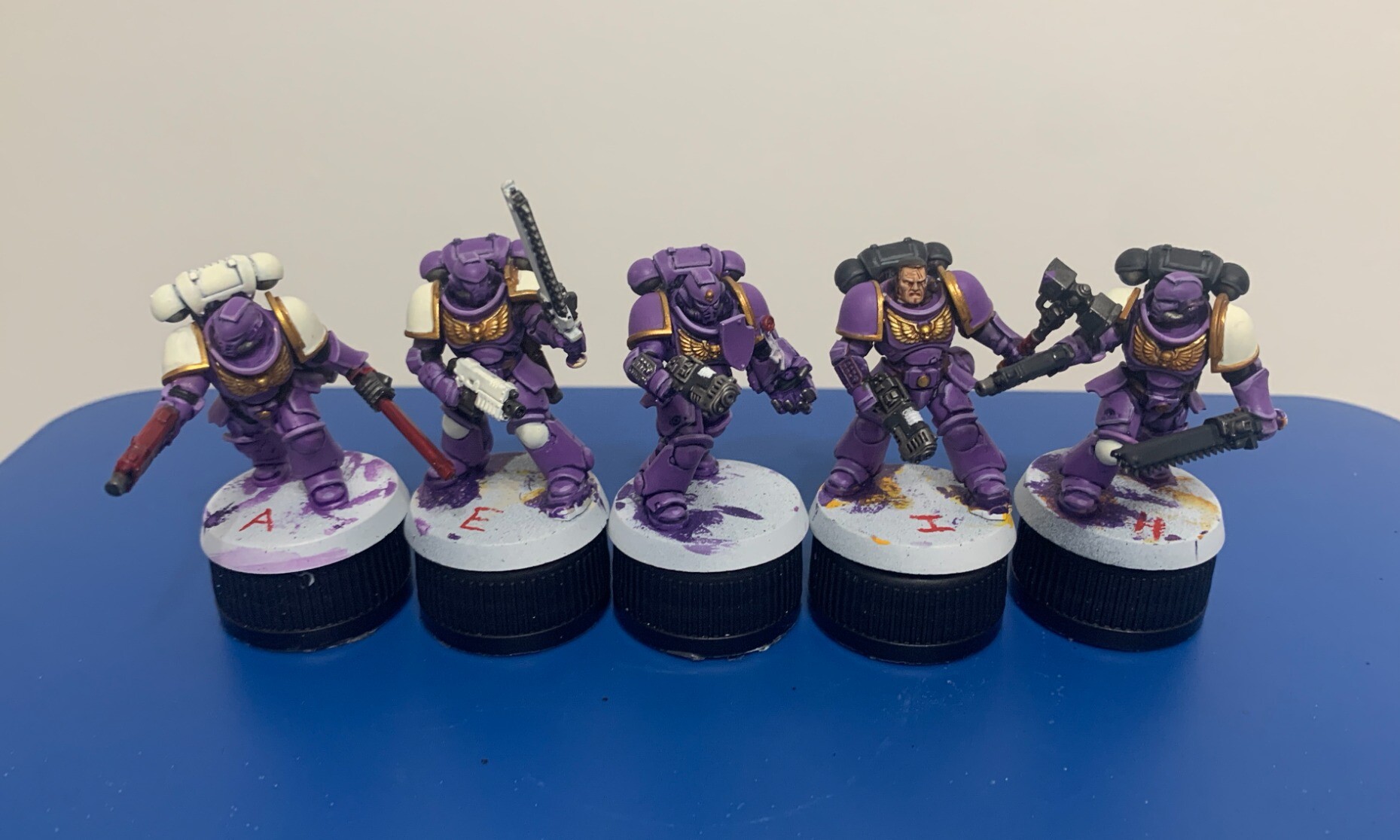 5 space purple space marines in a line with variations of accent colours. The purple is a middle brightness and appears warm, leaning more red than blue. The trim of the armour is gold. From left to right the variations are: White pauldrons with a white backpack and red weapons. Purple backpack, white pauldrons and white weapons. Purple backpack, purple pauldrons and back weapons. Black backpack, purple pauldrons and black weapons. And black backpack, purple pauldrons and black weapons.  The models are sat on black bottle caps on top of a blue plastic lid.