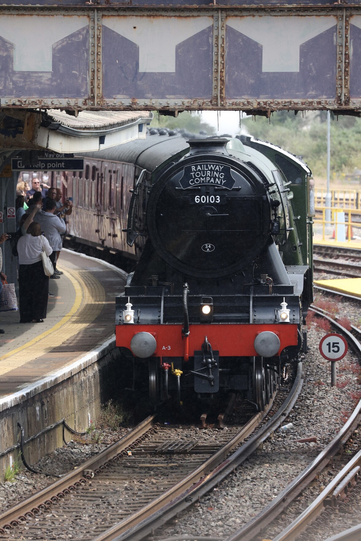 The front end of the Flying Scotsman at a train station. The Flying Scotsman is in dark green and black liver with a red front buffer block. To the right of the train there is a speed limit sign saying “15”. The platform has several people looking at and taking pictures of the train 