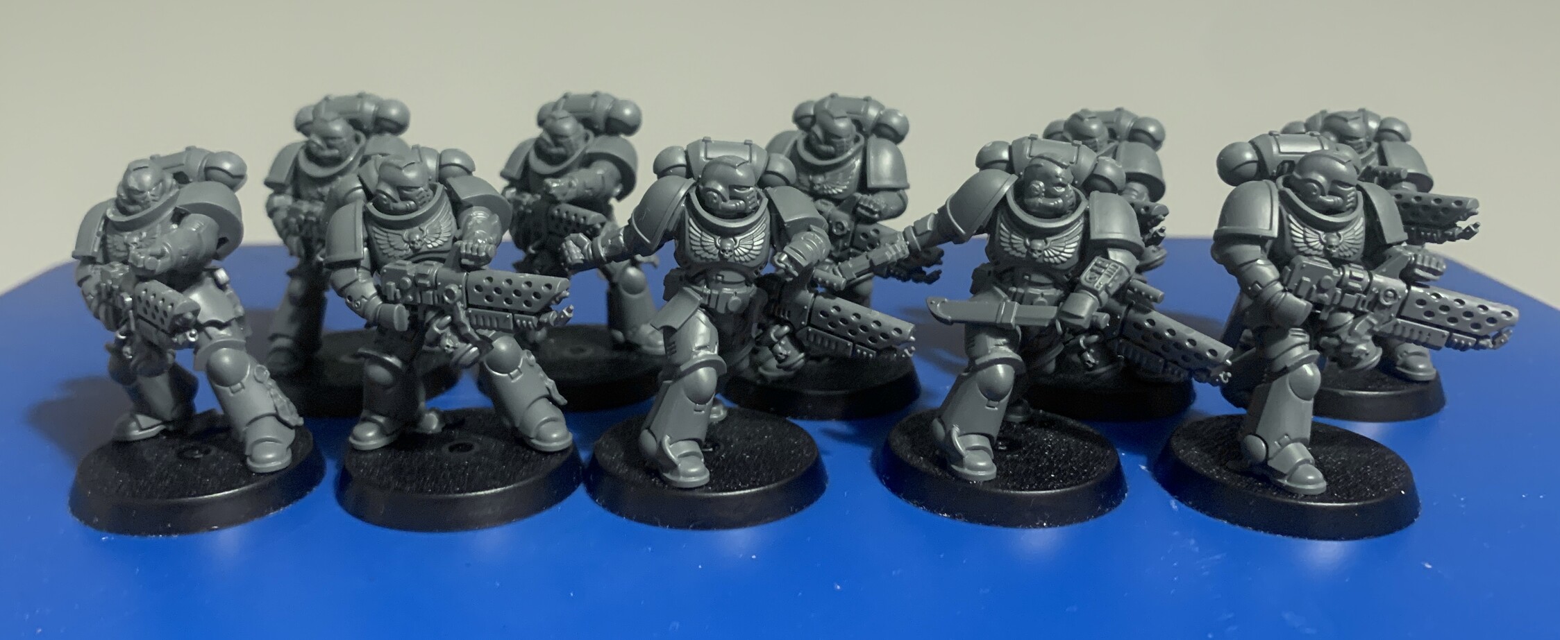 10 Infernus Marines from the Warhammer 40k universe stood in two ranks of 5. All models carry a pyre blaster which is a flame weapon that is shaped like a gun, has a pilot light at the front with the barrel protected by a perforated housing. The front center model has a pyre blaster on their back and is throwing a grenade. The model to the right of that also has a pyre laser on their back and is wielding a large knife and a pistol. The other 8 models are holding the pyre blaster in front of them with both hands in various positions.