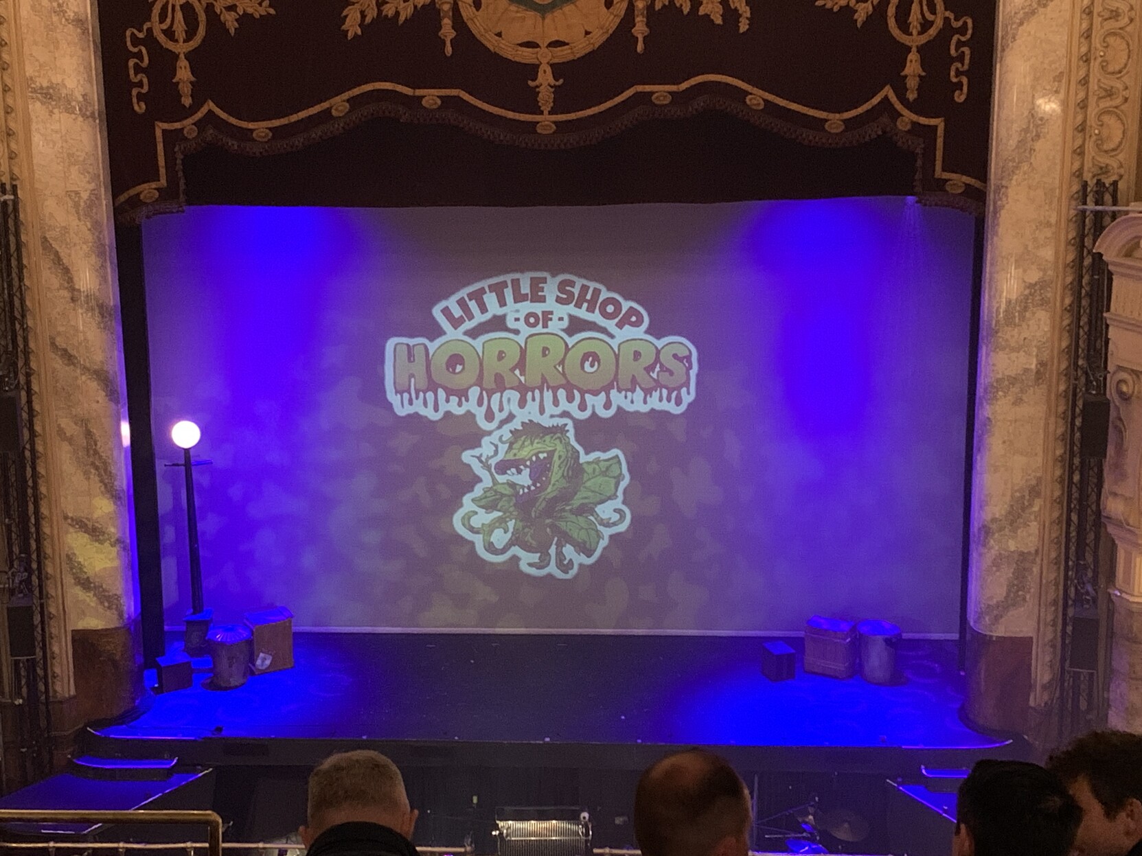 Looking down at a stage with blue lighting with the little shop of horrors logo. The stage is surrounded by while marble pillars
