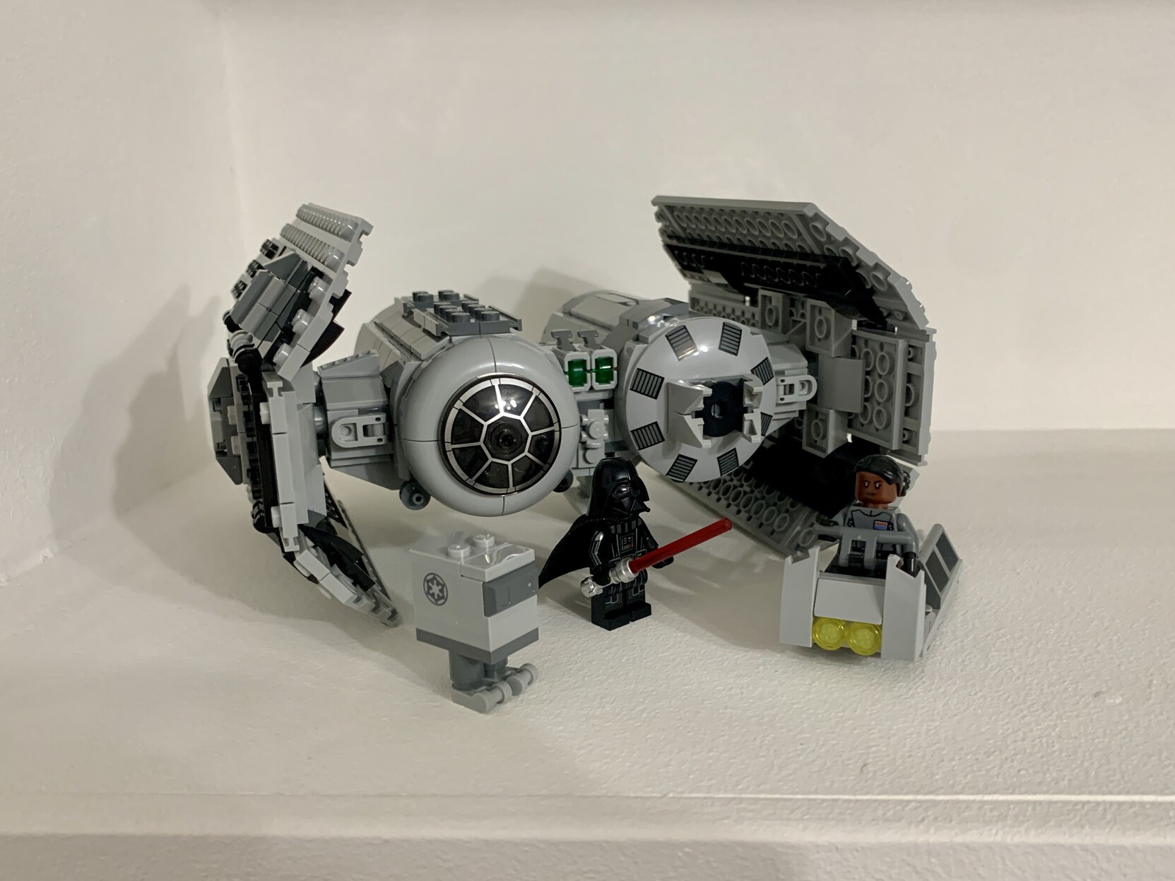 Lego Star Wars TIE bomber sat on a white shelf. In front of the bomber is an imperial gonk driod, Darth Vader with his lightsaber drawn, and vice Admiral Sloane piloting a trolley.
