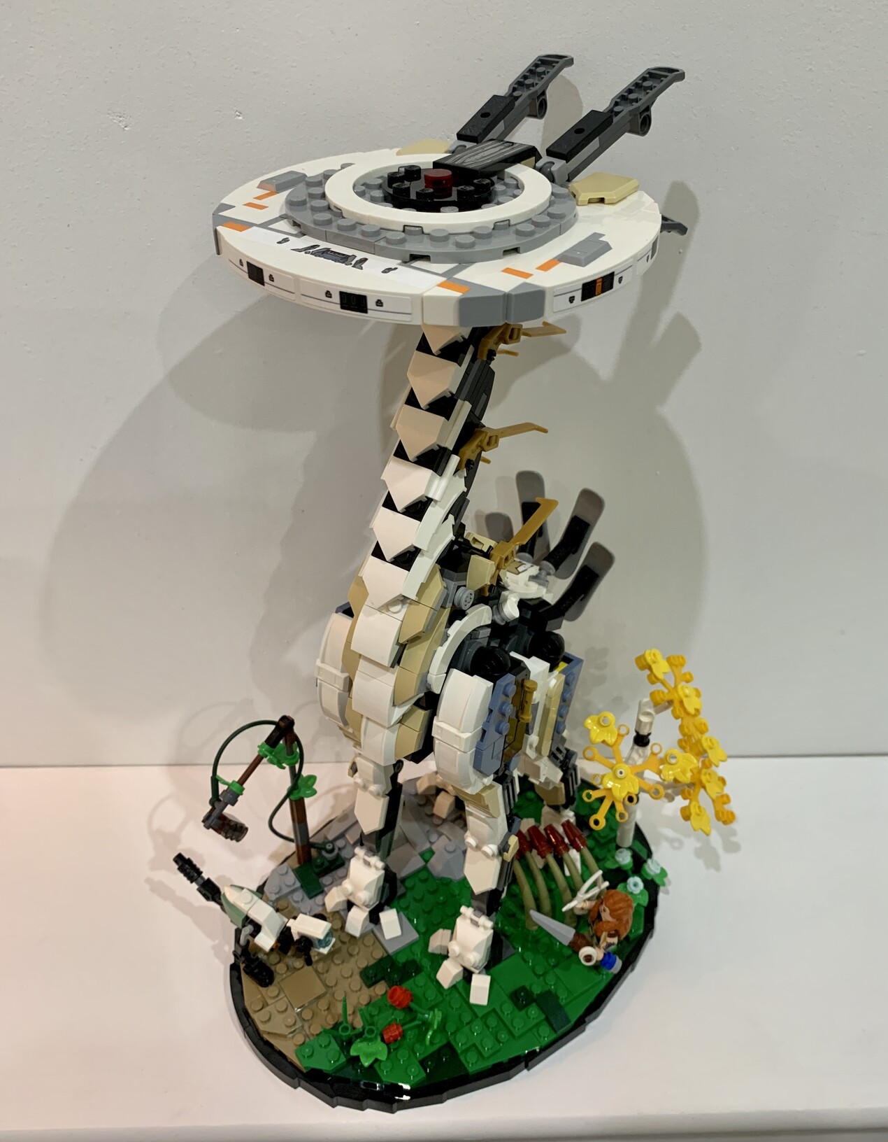Lego Tallneck from the Horizon Forbidden West game. It is shaped like a giraffe, but made of white, grey and black robotic like parts. It is stood on a grassy base with the character Aloy (human with red hair) and a small dog like robot