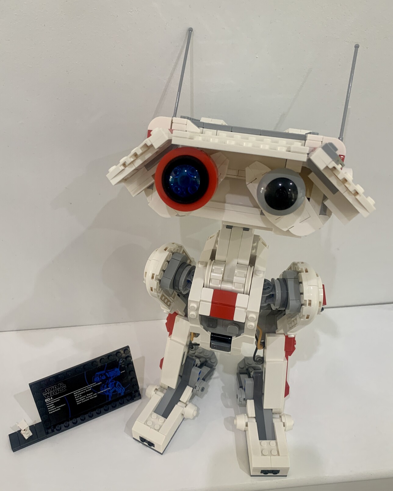 Lego BD-1. A short bipedal white, red and grey robot with an oversized head. The heads left “eye” is red while the right “eye” is grey. He has 2 antennas coming out each side of his head. His head is tiled up towards the camera