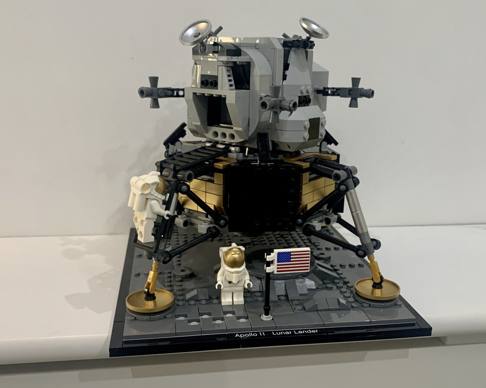 Built Lego Nasa Apollo 11 Lander. The Lunar lander is in a “just landed” configuration with all parts present. The lander is sat on a grey surface representing the moon. One astronaut is climing down the ladder on the closest left leg while the other is standing in front of the lander next to an American flag.
