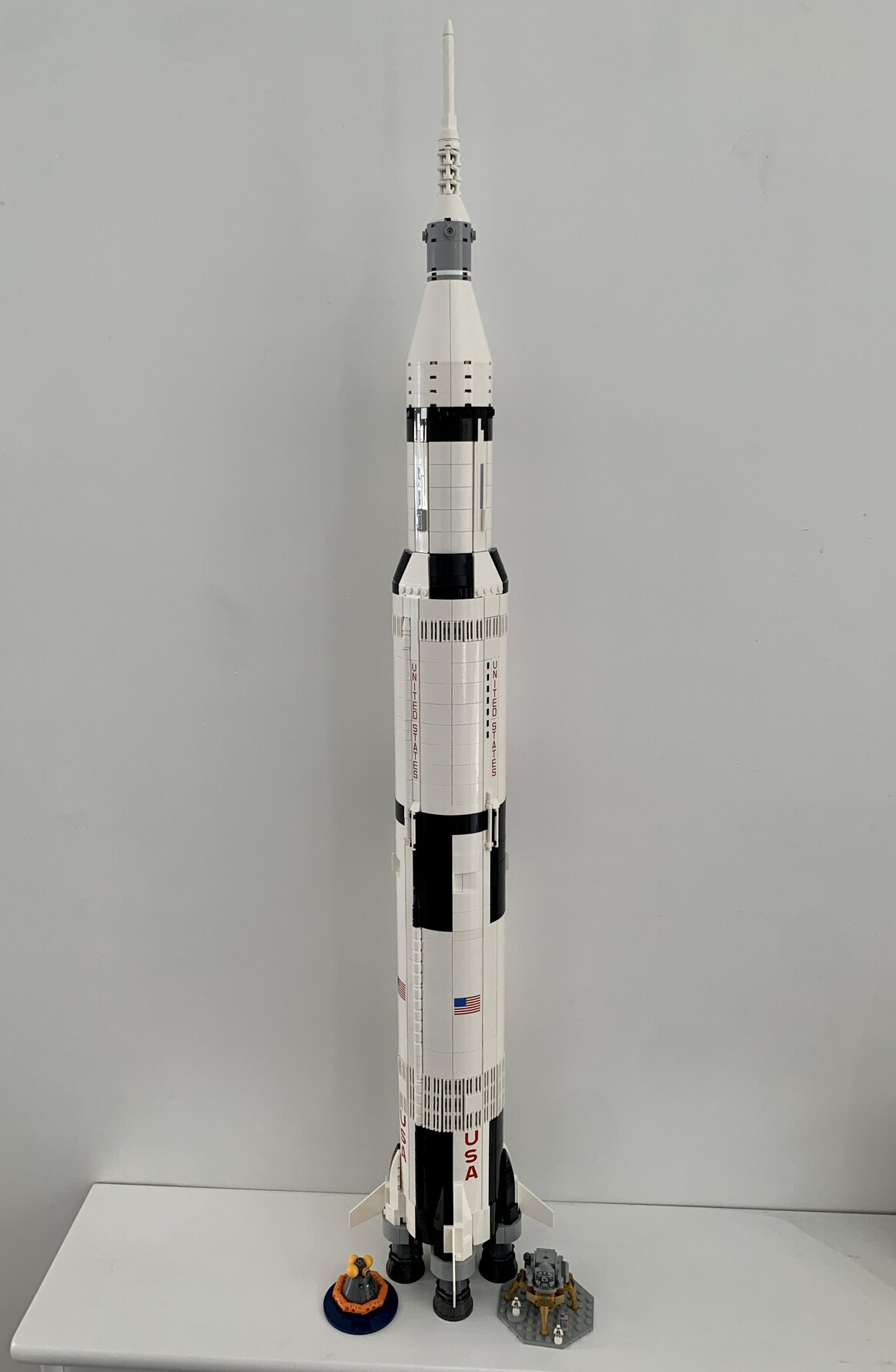 Lego Saturn V rocket standing vertically. To the right of base is a small diorama of the lunar lander one the moon with two astronauts stood in front of it next to a USA flag. To the left of the base is another diorama of the command module floating in the ocean with flotation equipment inflated.