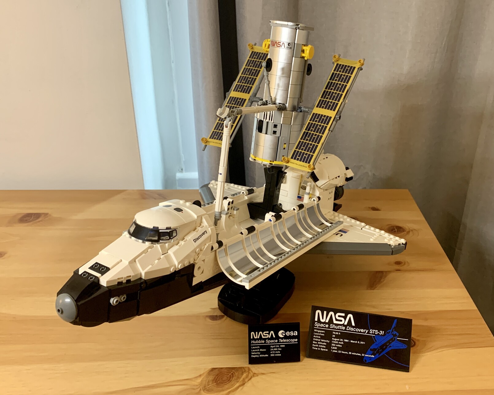 Lego Space Shuttle Discovery facing with its nose bottom left on a stand. Its cargo bay doors are open showing the Hubble space telescope above it. The Hubble is on a black stand and is pointing upwards. Its solar panels are deployed and it is connected to the Space Shuttleâ€™s manipulator arm. In front of the model are two plaques with details of the two space craft.