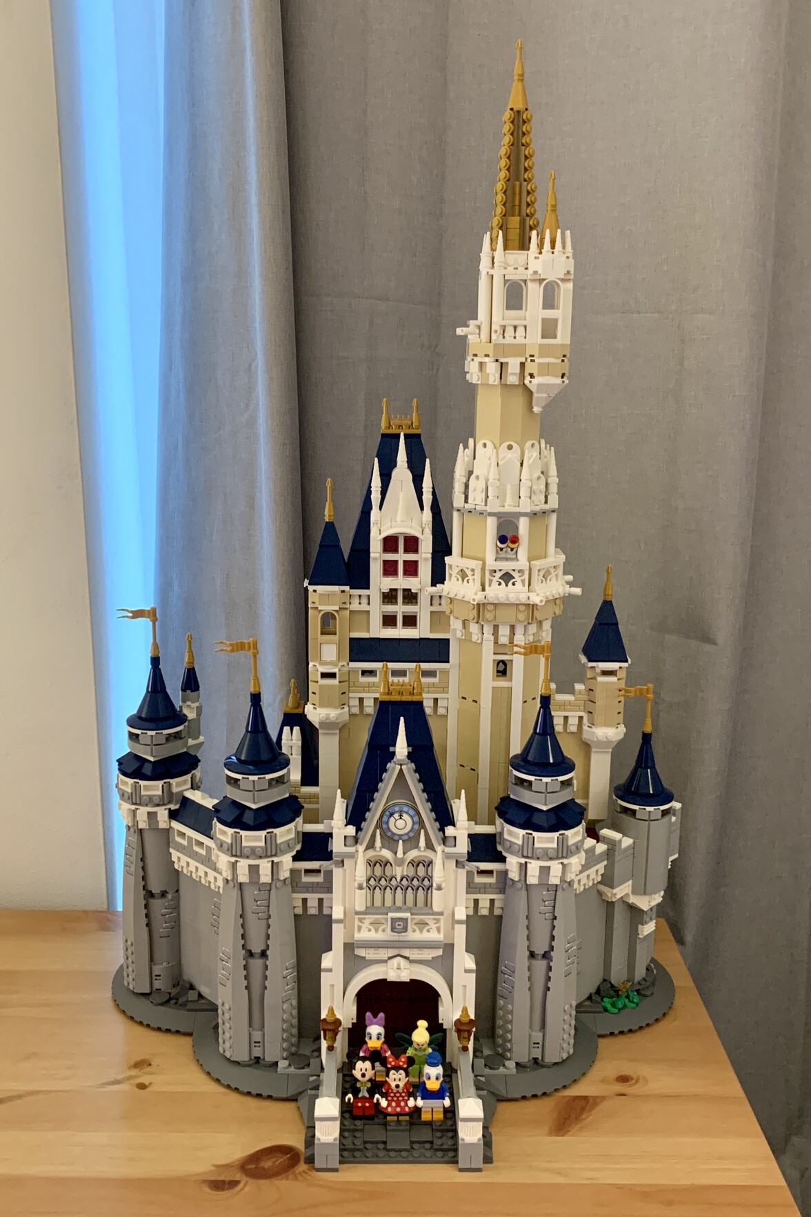 Built Lego Disney Castle (V1) sat on a brown desk with grey curtains behind. Mickey, Minnie, Donald, Daisy and Tinker Bell are stood infront of the main gates.