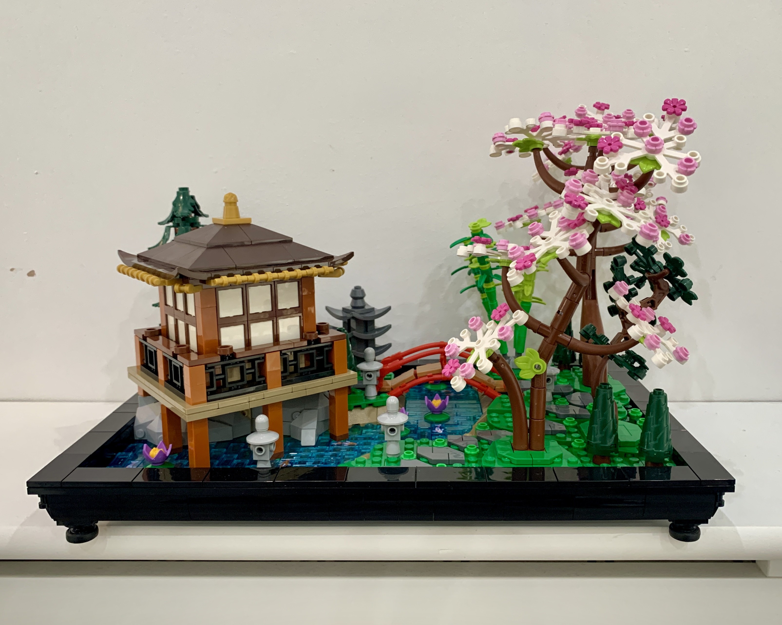 Built Lego tranquil garden. It is a Japanese-inspired garden scene with a wooden pavilion, red arched bridge, stream, koi carp tiles, lotus flowers, trees, rocks and lanterns. It is surrounded by a black border.