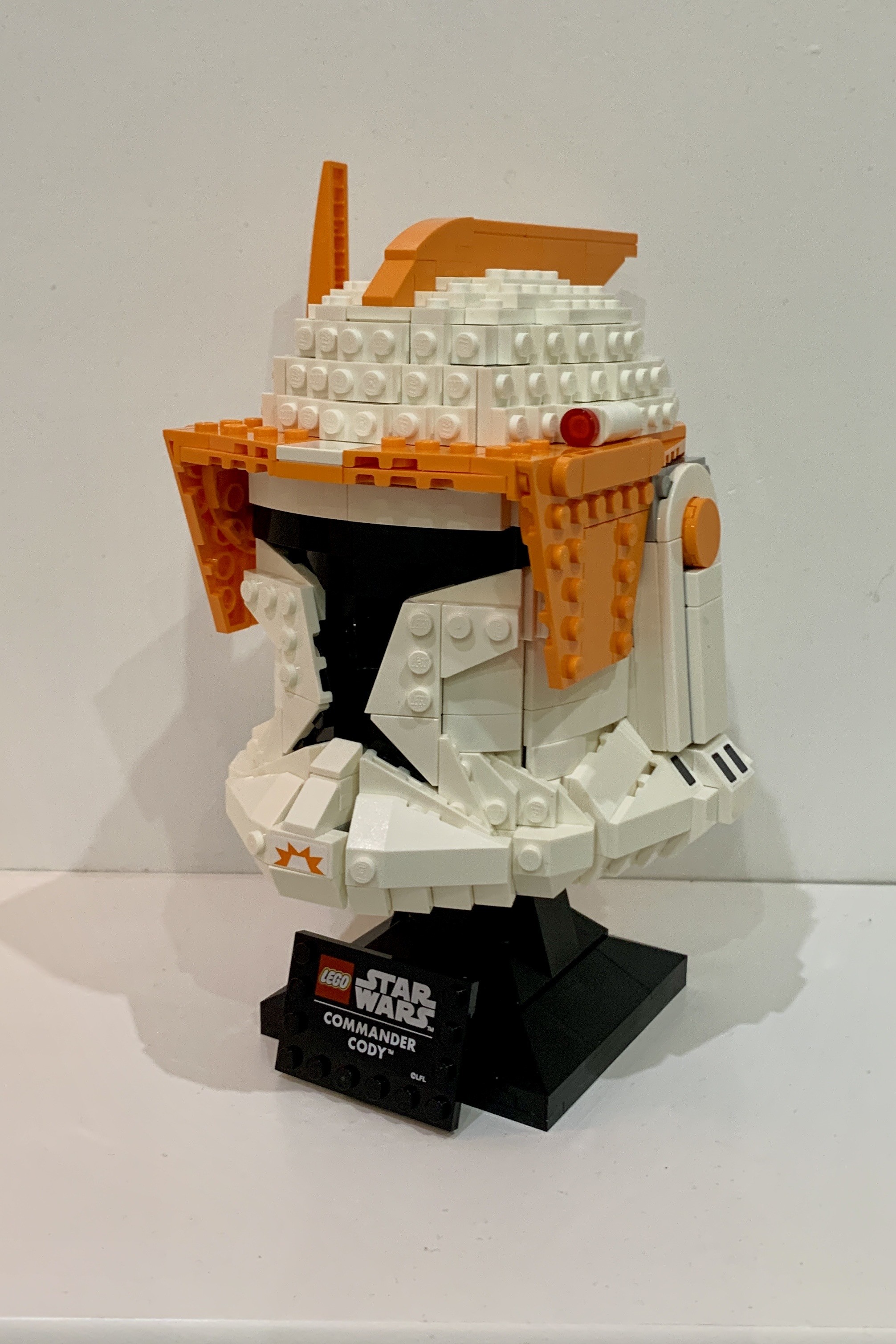 Lego model of Commander Cody’s helmet from the Clone Wars TV show. It is a white helmet with black visor. It has orange visor protectors,an orange antenna and a small orange fin on top of the helmet. It is on top of a black stand with a plaque that has the Lego StarWars combined logo and “Commander Cody™️”