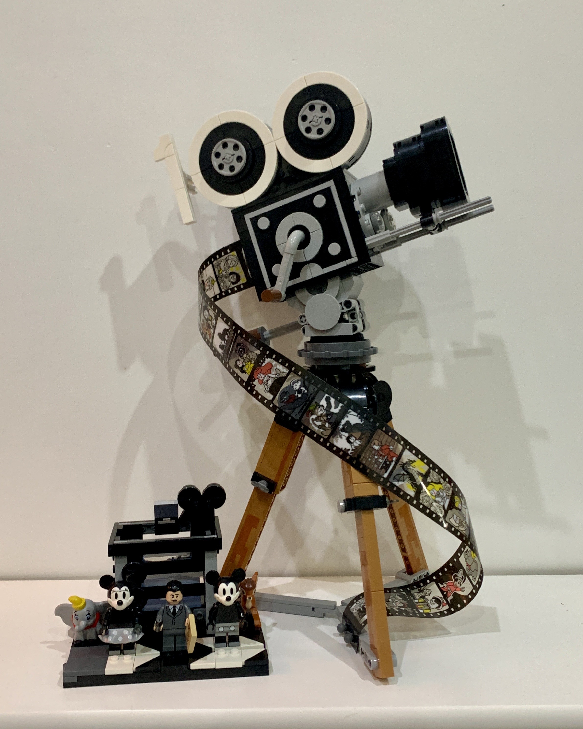 Lego Walt Disney Tribute Camera. It is an old fashioned film camera on a wooden tripod. The reels on top of the camera make two 0’s, to the left of them is a 1 so the top of the camera reads 100. Coming out of the back of the camera and wrapping around the legs is a film strip featuring frames from many of Disney films. To the left of the camera and tripod is a small model of a multiplane camera. In front of that are 5 minifigures representing Walt Disney, Mickey and Minnie Mouse in black and white, Dumbo and Bambi. The minifigures and multiplane camera are sat on a black and white clapperboard as a base.