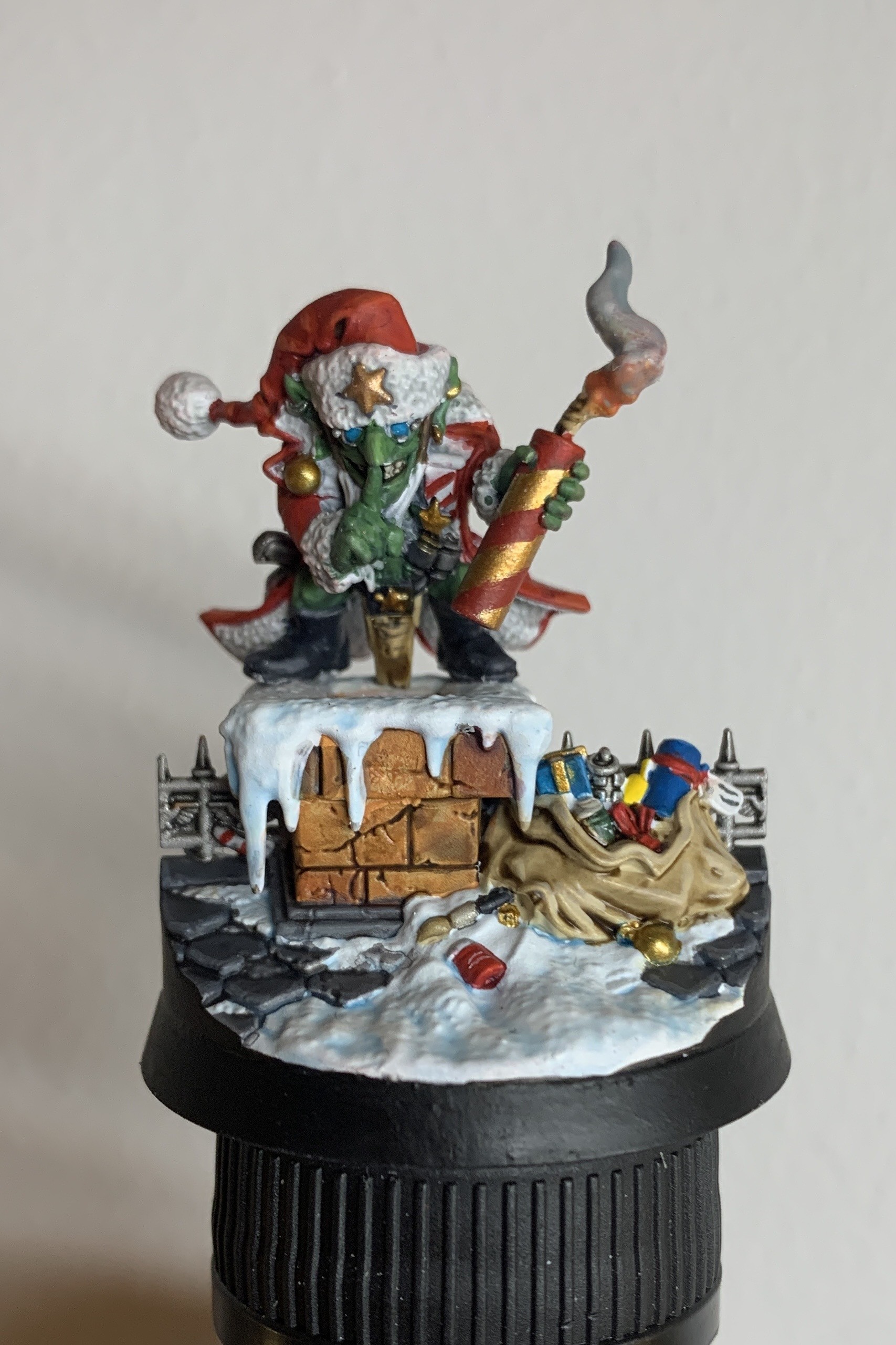 Small green Oak in a red Christmas outfit hiding a red and gold explosive. The Ork is stood over a chimney and is about to drop the explosive down it. Next to the chimney is roof tiling and a sack of presents