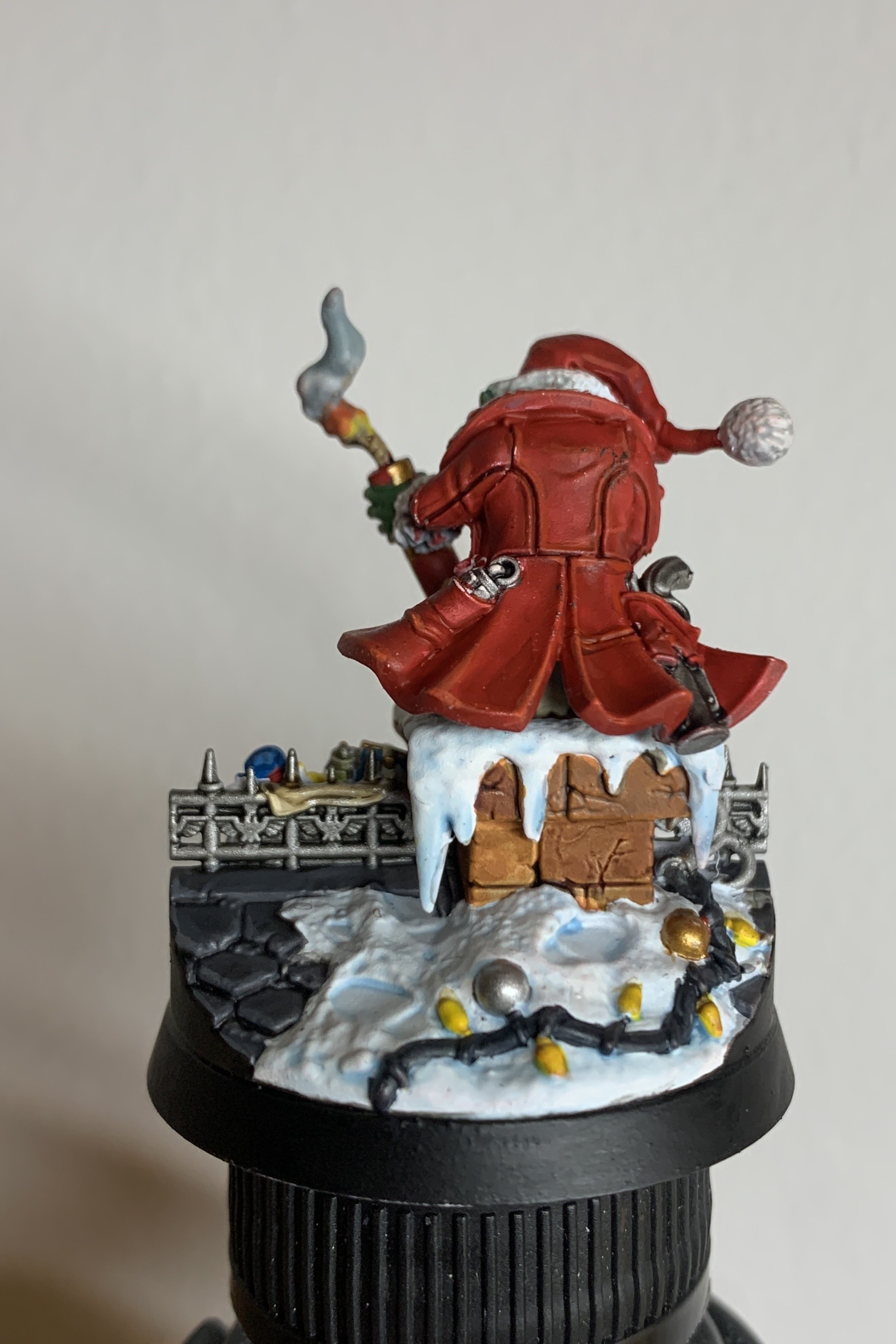 Back of an Ork in a red Christmas outfit standing over a chimey. The Oak is holding an explosive. Next to the chimney is roof slates and a throwing line with an hook made out of a string of Christmas lights