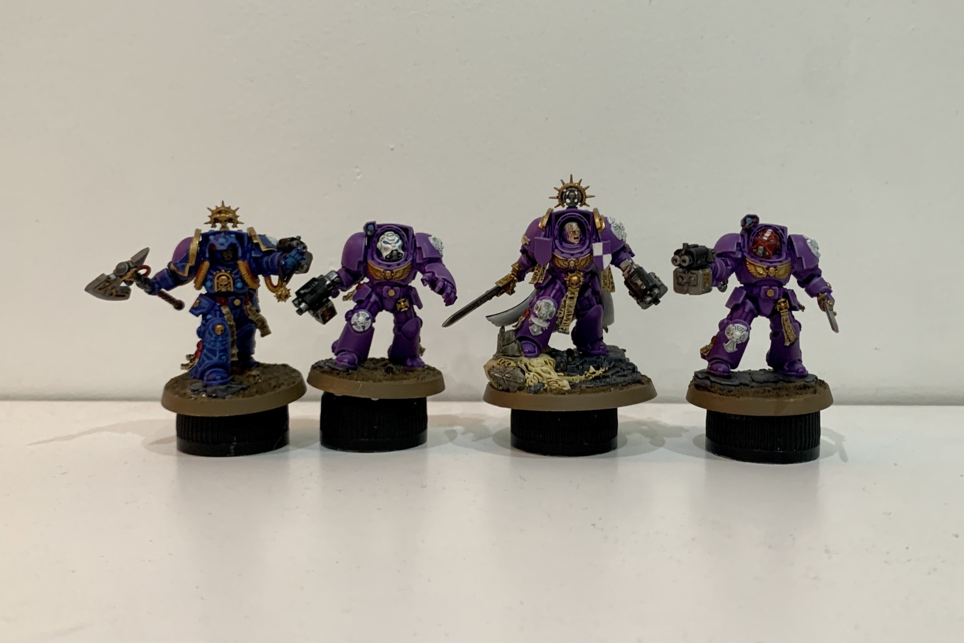 Four Warhammer 40k Terminator models. The leftmost is a Librarian in dark blue armour with gold trim, in his right hand is a ax, his left hand is held upwards and glowing blue. His armour also has several mutlicololured tubes. The other three models are in purple and gold armour. The righ centre model of these three is a captain stood on a the corpse of a Tyranid. The rightmost model is wearing a red helmet with a white stripe and the other model has a white helmet.