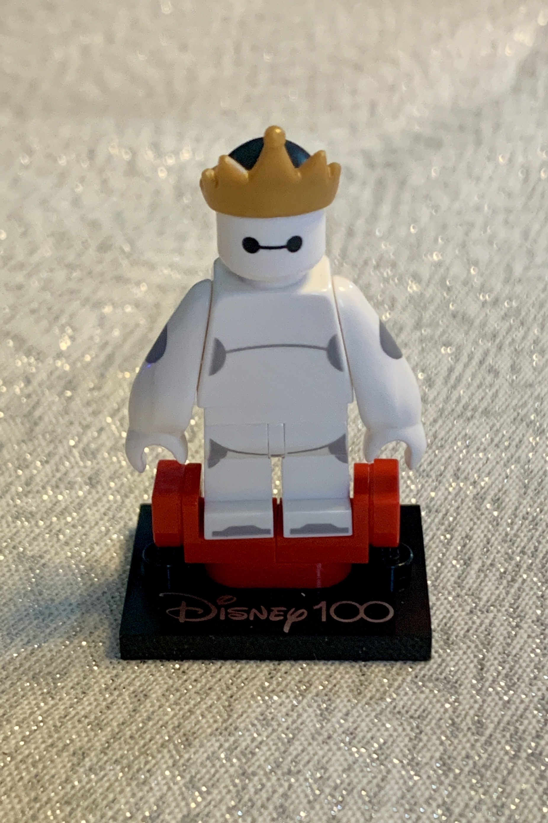 Lego Baymax minfigure wearing a gold crown, stood on a red stand