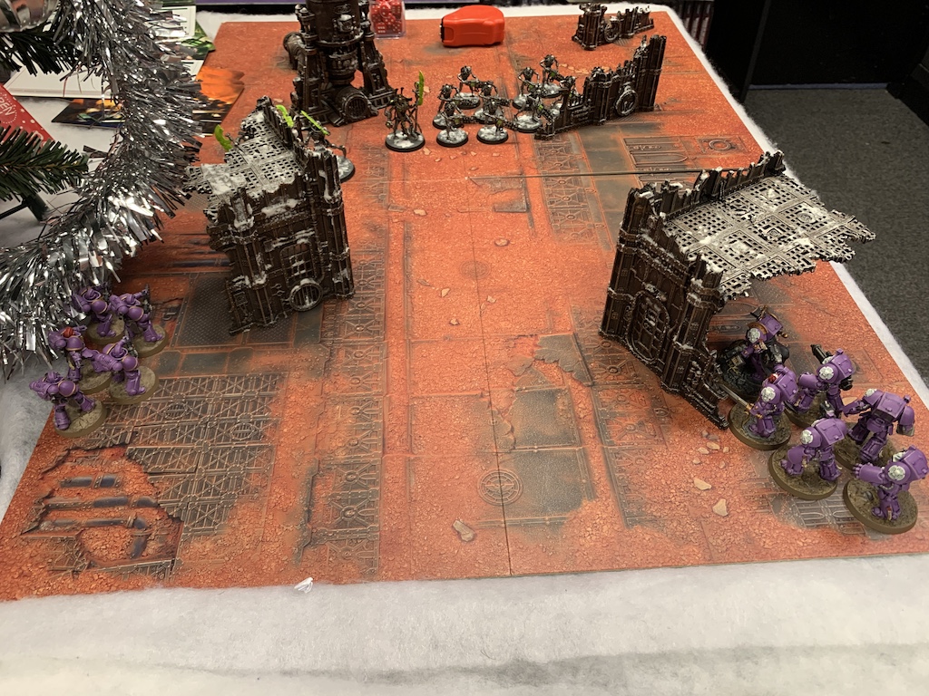A battlefield with various bits of terrain. In the bottom right corner is six purple Space Marines in Terminator Armour hiding behind a ruin. At the bottom left are five purple Infernus Marines hiding behind a ruin. In the centre are ten Necron Warriors and a Necron Overlord