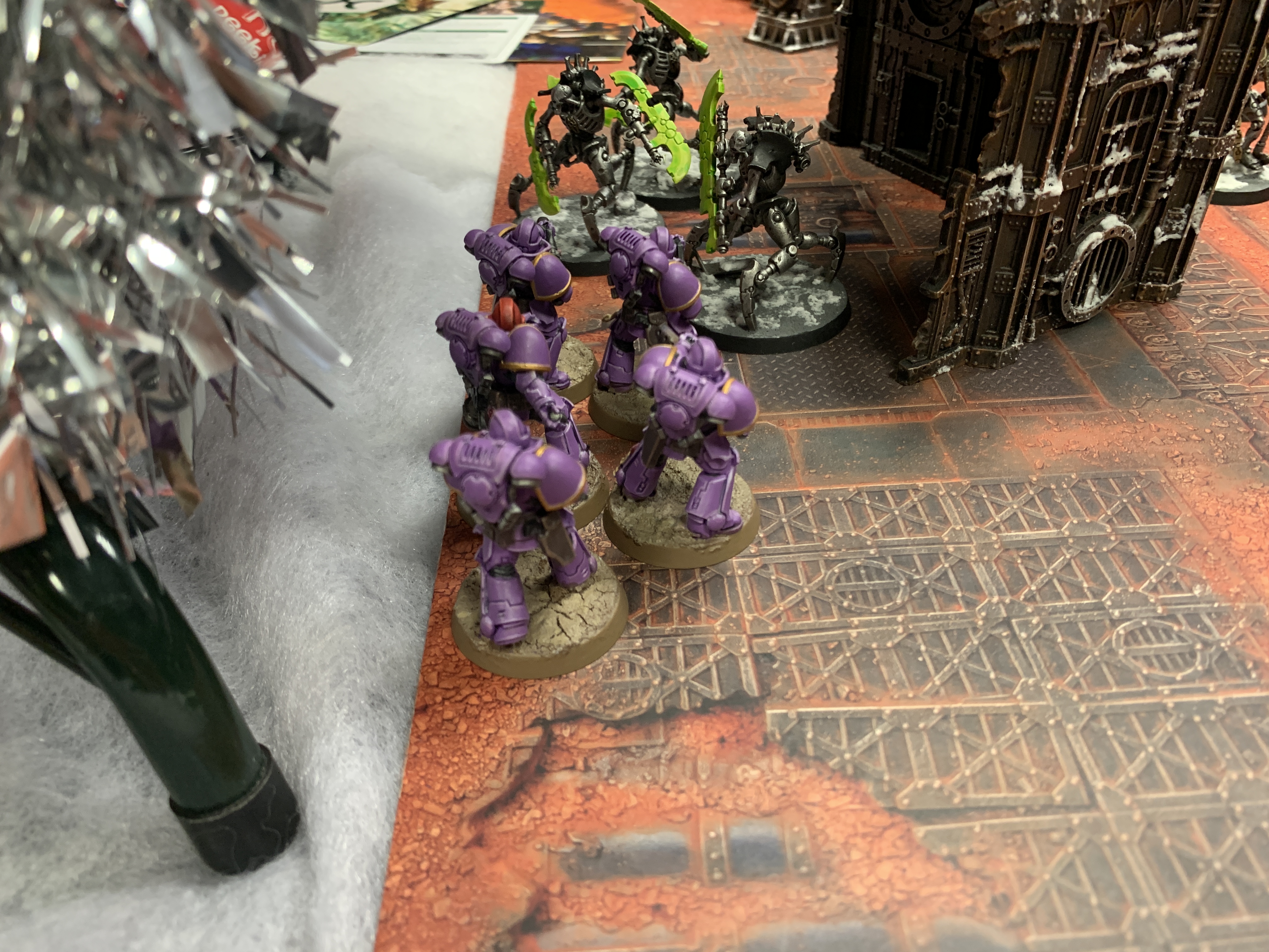 Five purple Infernus Marines facing three Necron Skorpekh Destroyers wielding light green blades. They are stood next to a bit of scenery and some tinsel from a Christmas tree