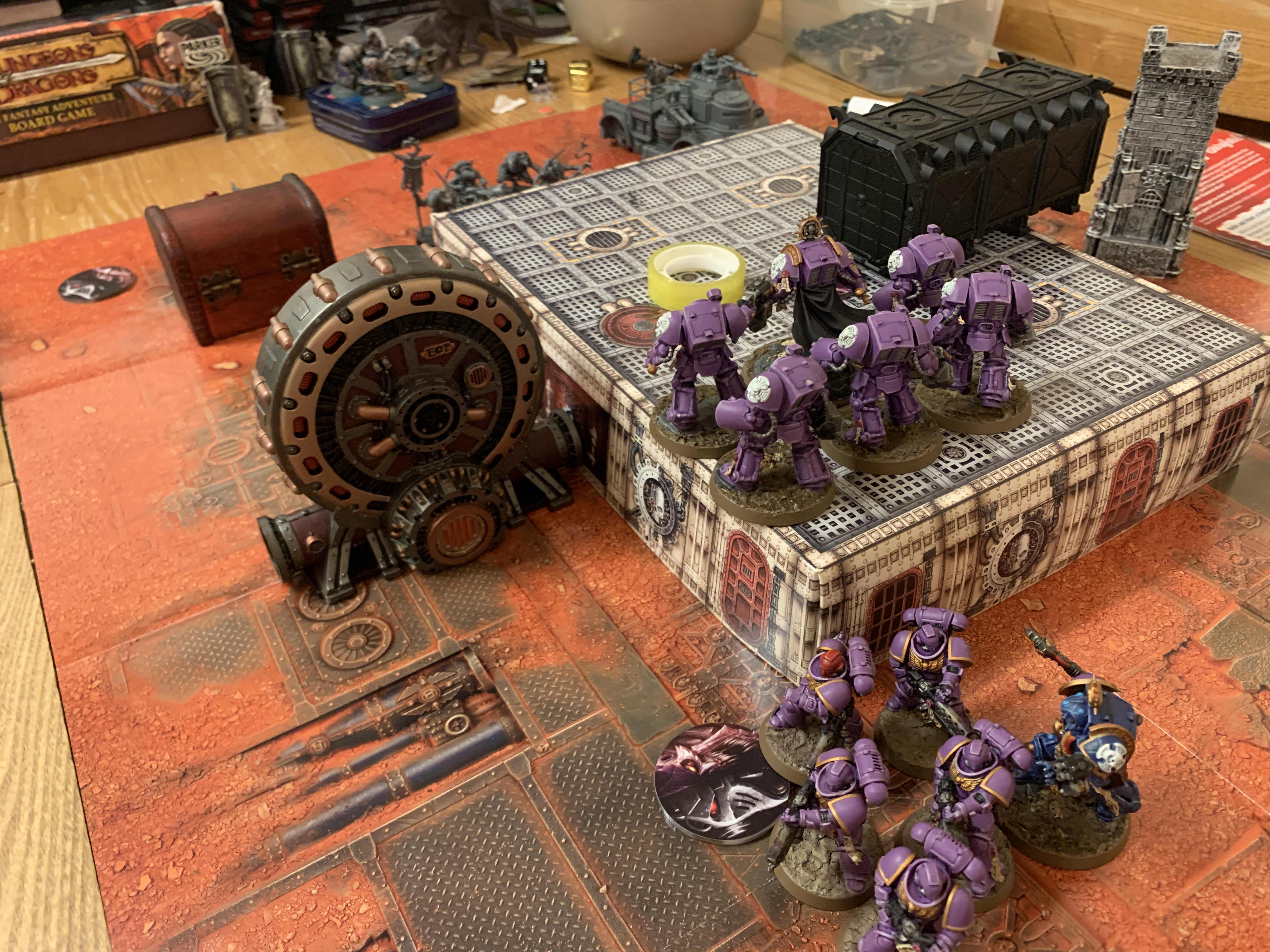 A battlefield split in the middle by a boxy bit of terrain that splits the area. On the lower area behind the terrain is a Genestealer truck and 5 Acolytes. On the terrain is six purple Space Marines in Terminator Armour. At the front on the lower level are 5 Infernus Marines in purple armour and a Librarian in dark blue armour. The battle field has several other terrain features, including some boxes, creates and a circular power generator
