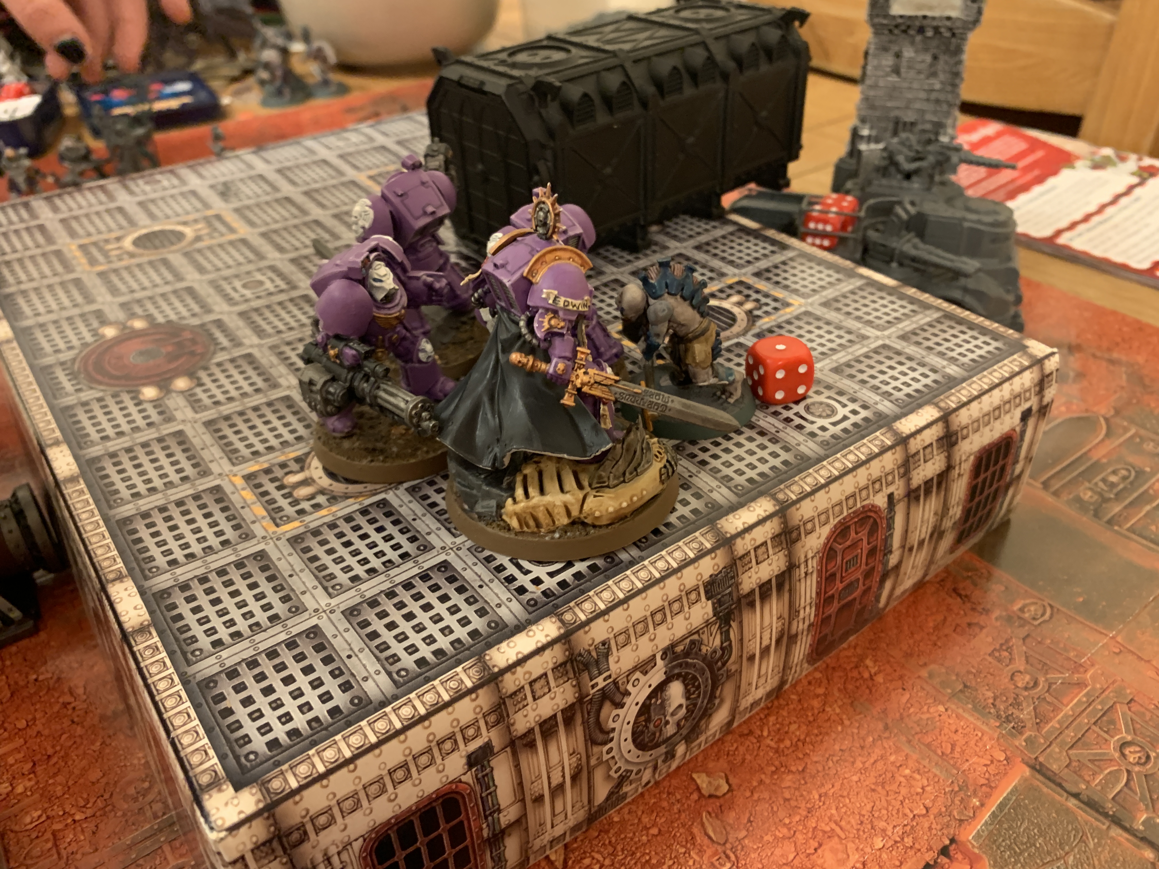 Four purple Space Marines stood on a boxy bit of terrain facing a single Aberrant. Off to the side of the terrain is an unpainted truck. In the background is a large group of Neophyte Hybrids