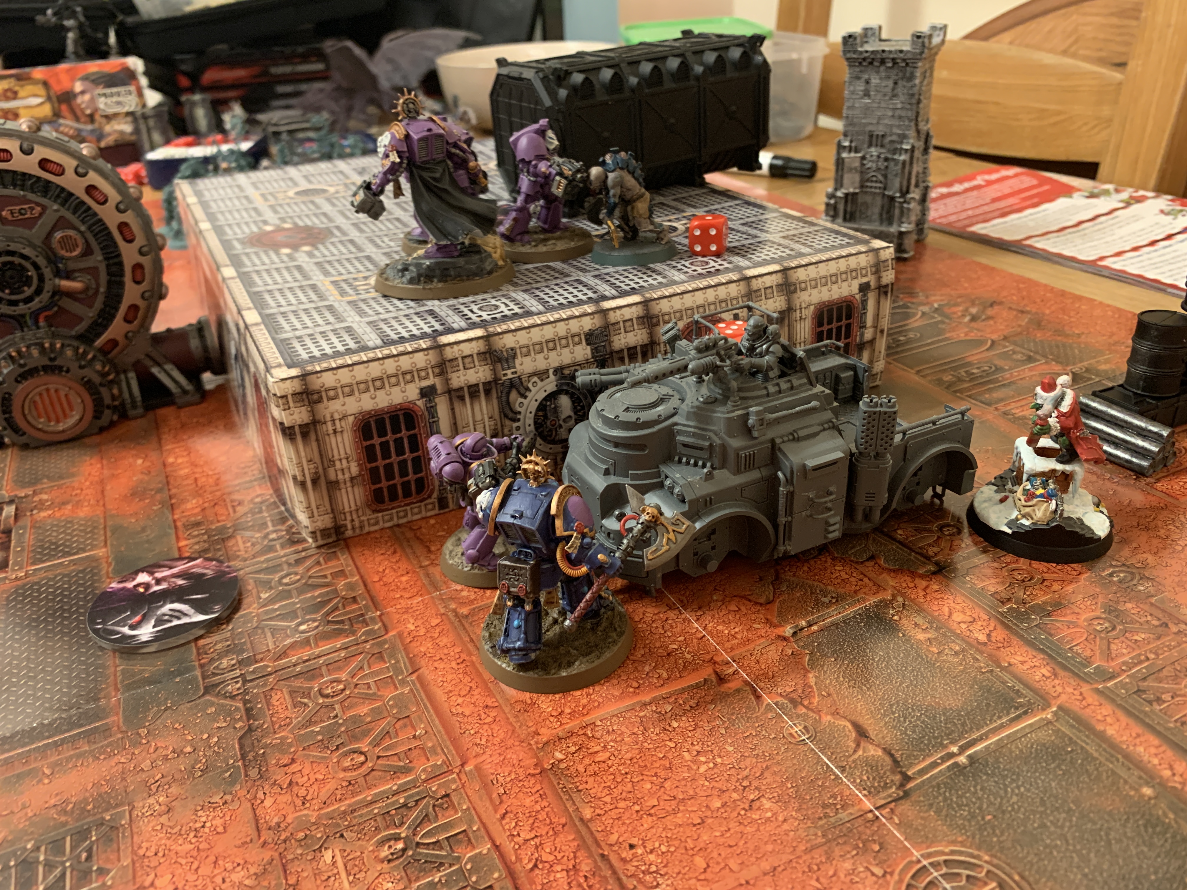 A battle scene split into two levels by a boxy bit of terrain. On top of the terrain are three purple Space Marines in Terminator Armour fighting a Genestealer Aberrant with a red dice next to it showing one. On the lower level is a unpainted truck with a purple Infernus Marine, a dark blue Librarian in Terminator Amour and a Ork in a Santa costume on a snowy base 