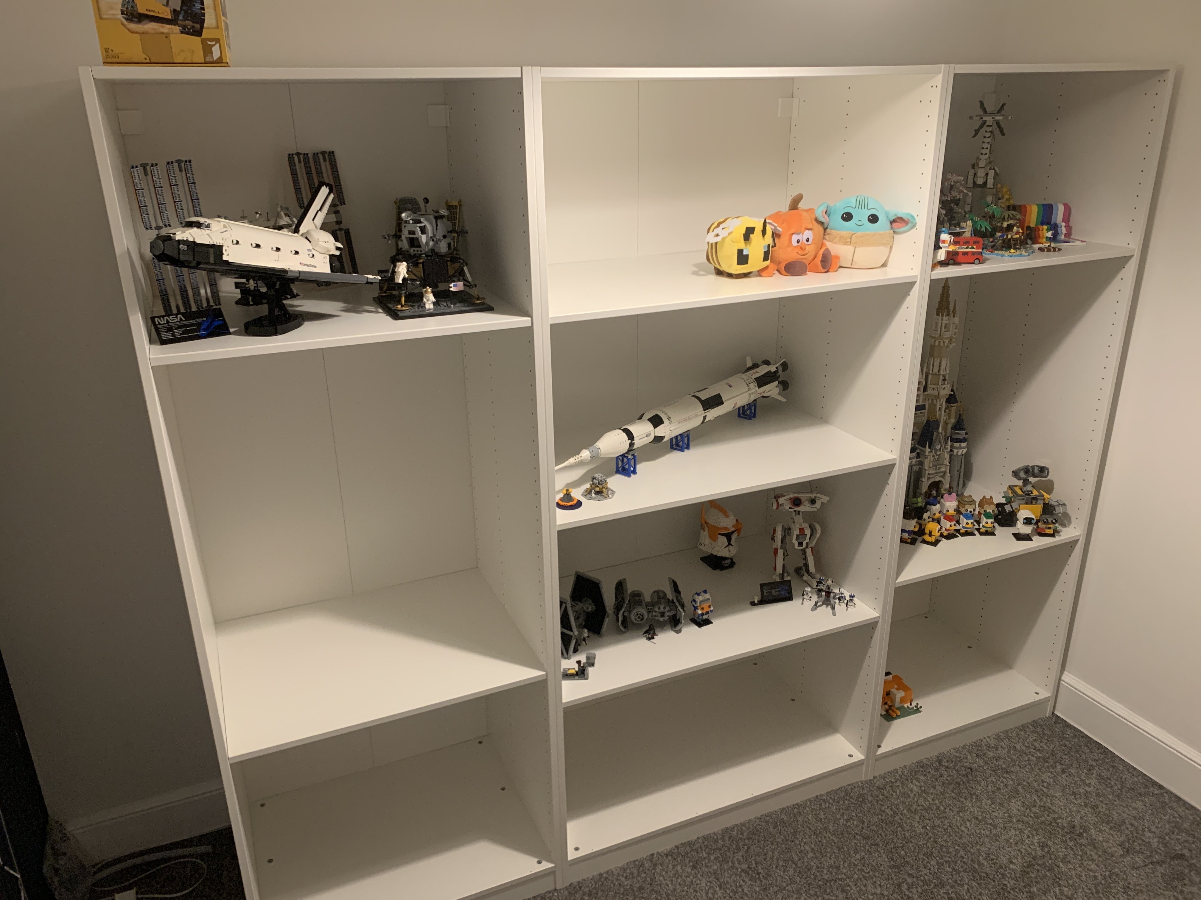 Three white shelving units stood next to each other. On the shelves are various Lego sets and some plushies. Top left is a collection of NASA models, including the ISS, Space Shutter and Lunar Lander. The top centre has three plushies: a Minecraft bee, a pumpkin with eyes, and a Grogu. Below that is a model of the Saturn V rocket. Below that is a collection of Star Wars models, including a TIE fighter and bomber. BD-1, a Clone Trooper helmet and a small model of Ahsoka. The top right is a mixed collection of models, including a red London bus, a duck, a Tallneck, and a topical island scene, and the Everyone is Awesome set features a rainbow resign. Below that is a large Disney castle, WALL-E and several assorted Disney characters. Below that is a Minecraft Fox house