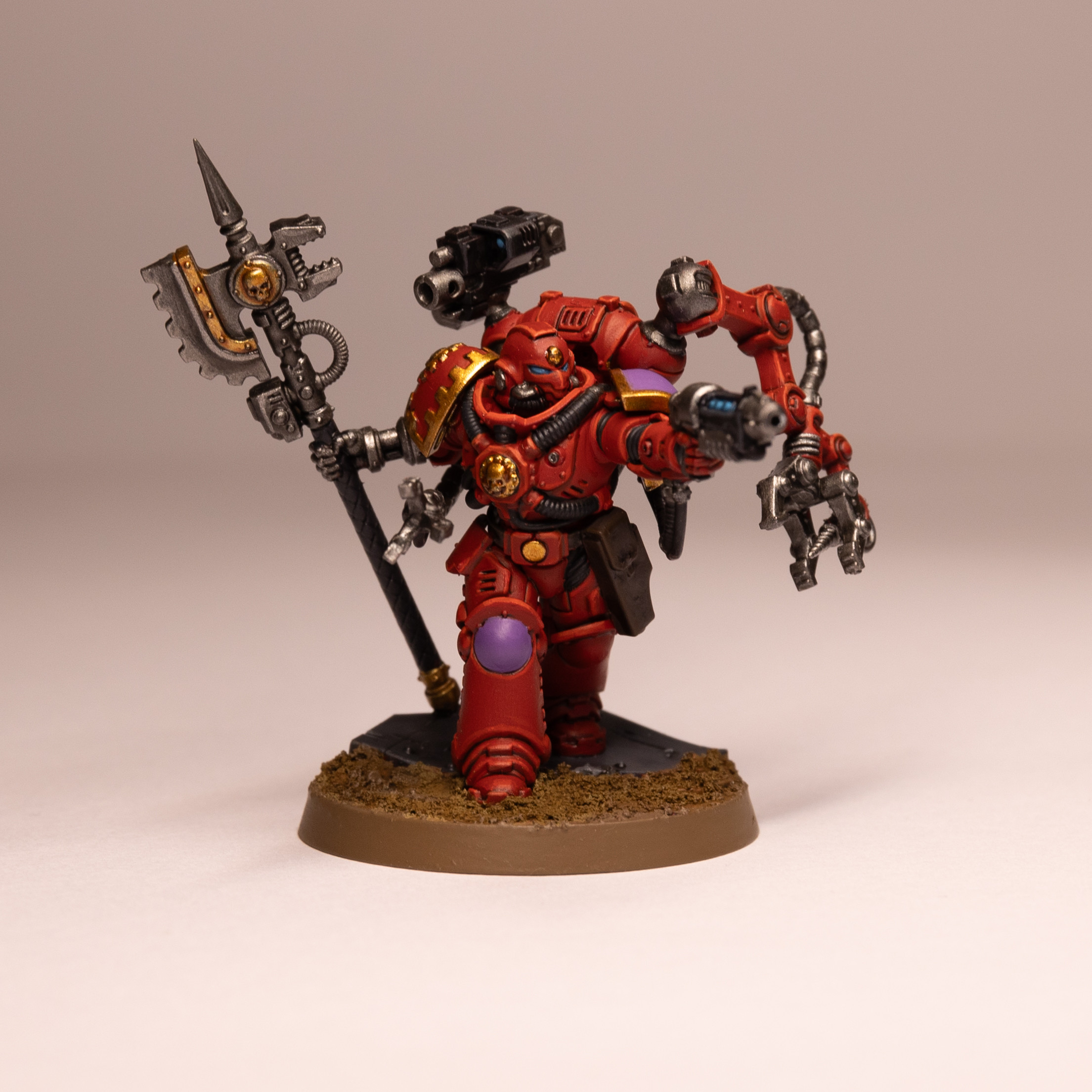 Red Space Marine Techmarine with a purple shoulder and knee pad, stood on a sandy coloured base. The Techmarine has a robotic right hand holding an elaborate axe, above their right shoulder is a backpack mounted gun. In their left hand they’re holding a gun with a glowing blue barrel and on the left side of their backpack is a robotic arm with a claw and drill at the end. The Techmarine is looking to the right of the image looking down the gun sights.