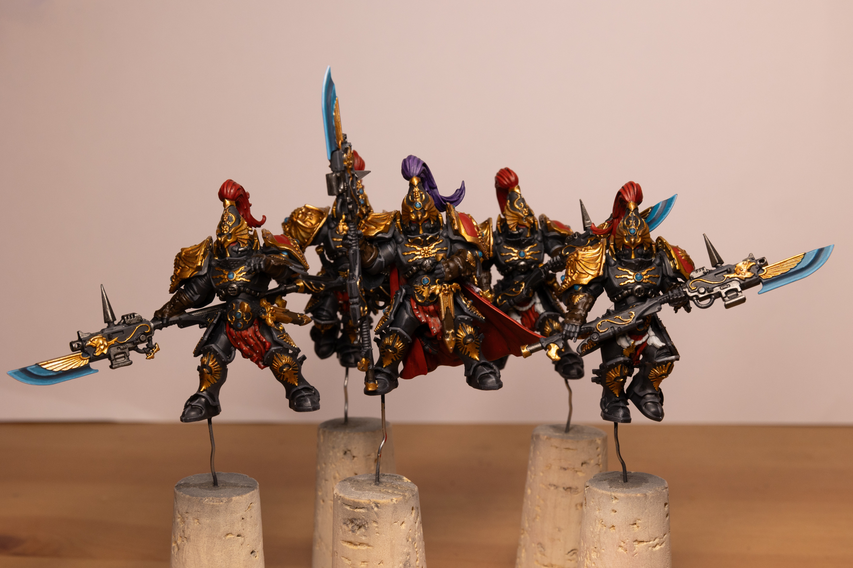 Five Warhammer 40k Custodian Guard in the Shadowkeepers color scheme. They are five tall armoured people in black armour with gold trim and red accents. They have tall helmets with plumes of hair, they are all red except the Captain in the middle who has a purple plume. They are all carrying spear weapons whose tips are an electric blue. All the models are mounted on metal sticks and corks.