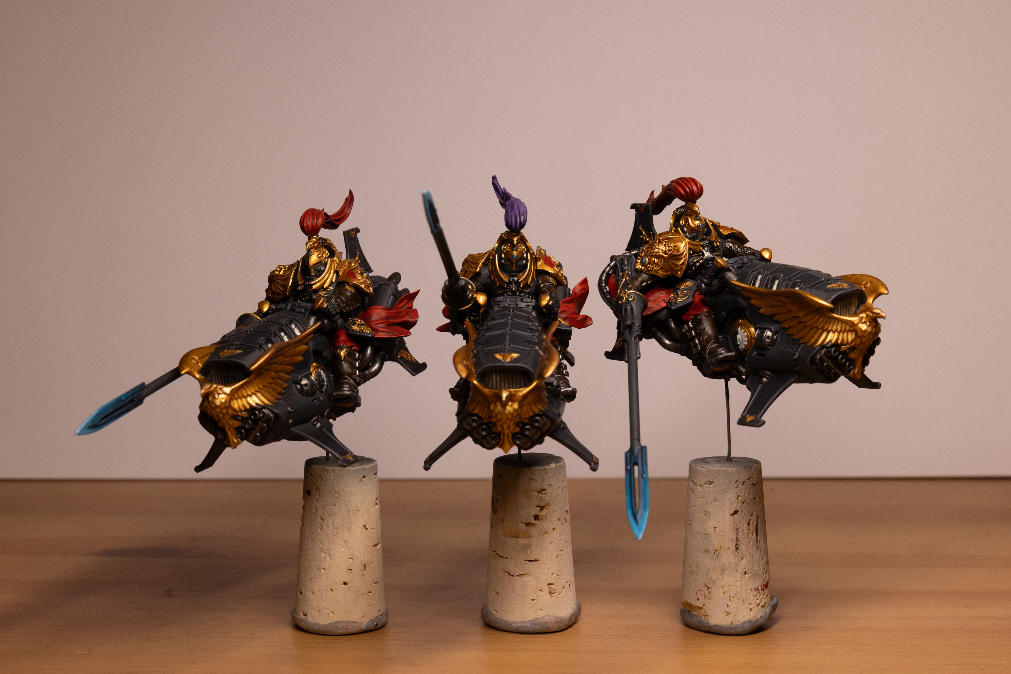 Three Warhammer 40k Custodian Vertus Praetors in the Shadowkeepers color scheme. They are three armoured warriors on large jet bikes painted in black armour with gold gold trim and red accents. The front of each bike features a large golden eagle and each warrior is holding a long lance with a glowing electric blue tip. The central warrior has a purple head plume. All three models are mounted on metal sicks and corks.