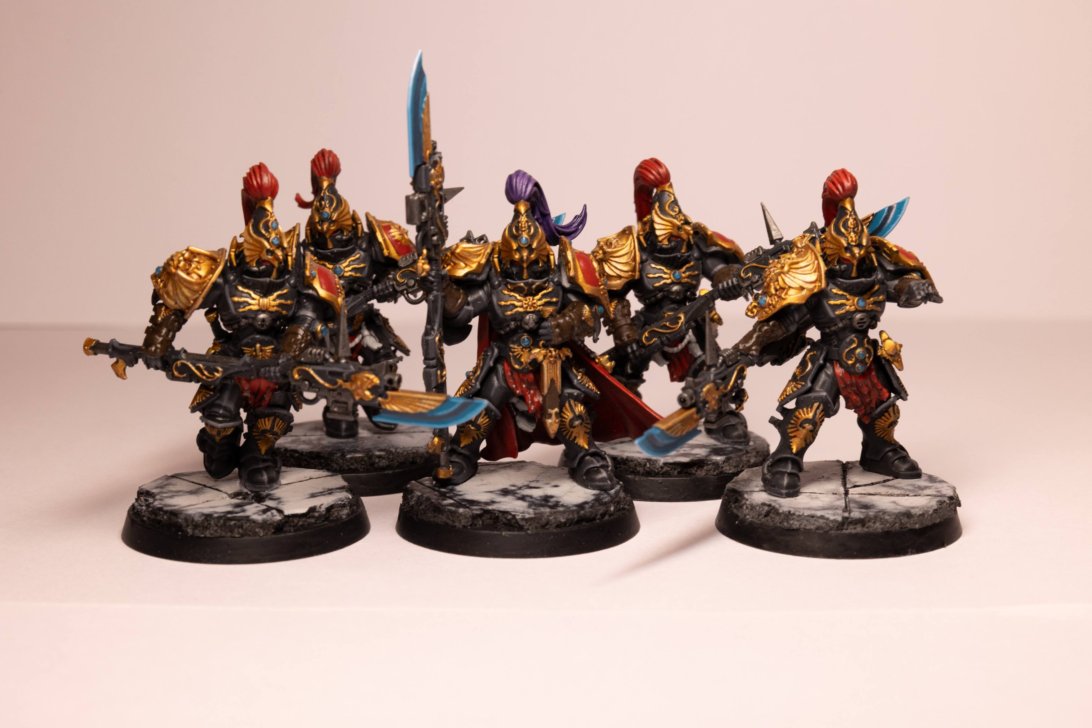 Five Warhammer 40k Custodian Guard in the Shadowkeepers color scheme. They are five tall armoured people in black armour with gold trim and red accents. They have tall helmets with plumes of hair, they are all red except the Captain in the middle who has a purple plume. They are all carrying spear weapons whose tips are an electric blue. They are all stood on white and black marble-like bases.