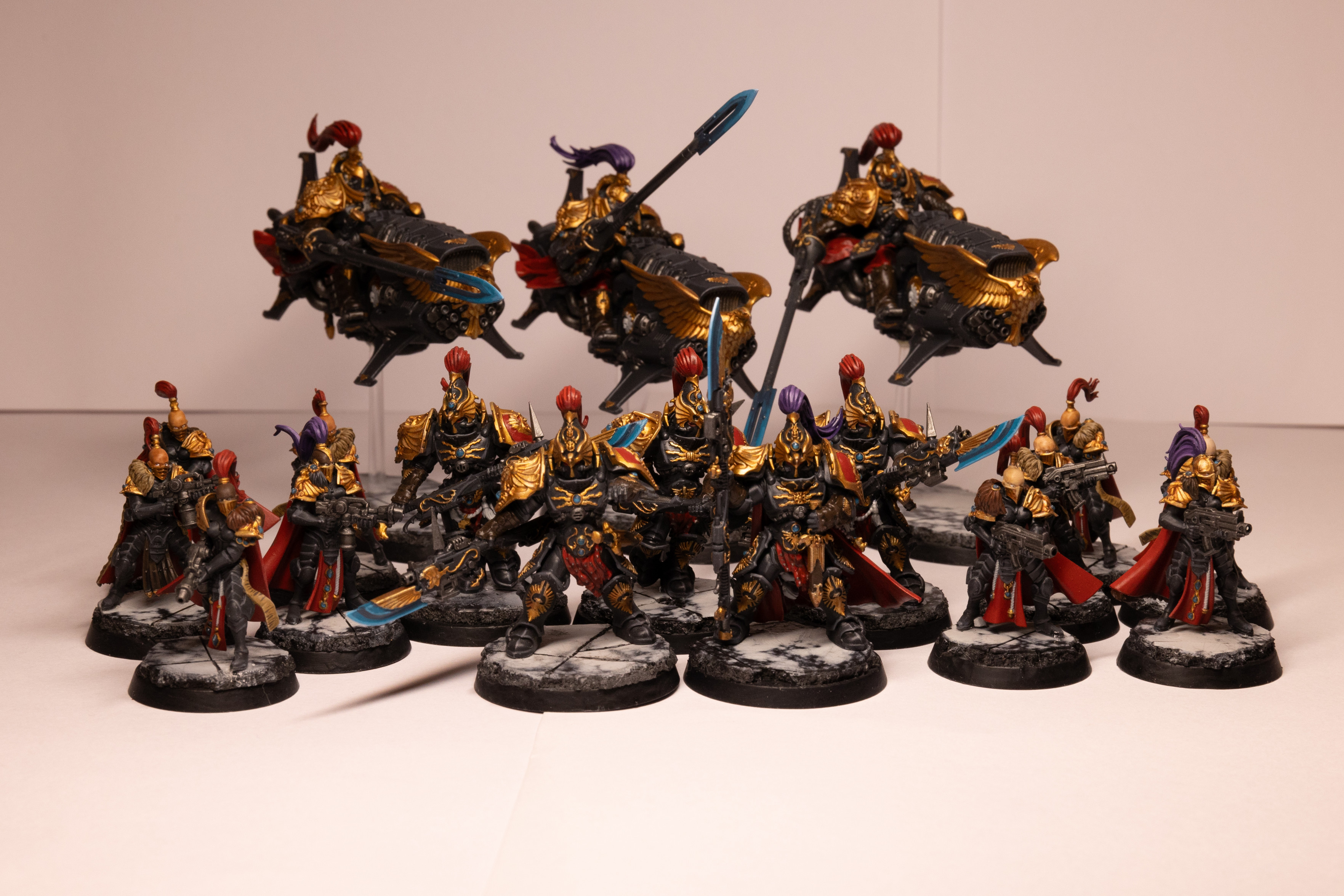 Small Warhammer 40k Custodes force in the Shadowkeepers colour scheme. In the front are five Custodian Guard, large armoured warriors in black armour with gold trim and red accents and head plume. They are weilding large spear like weapons with glowing blue tips. Either side are them are two groups of five Sisters of Silence, sleekly armoured warriors in black and gold, with red accents. They are holding either a flame weapon or bolt gun. They are either bald and a top knot red plume, or have a gold helmet with head plume. At the back are three Vertus Praetors. They are three large jet bike vechiles each piloted by a warrior in large armour. They are black and gold with red accents and head plumes. On the front of each bike is a large golden eagle. Each warrior carries a long lance like weapon with a glowing blue tip. All models are mounted to a black and while marble-like base.