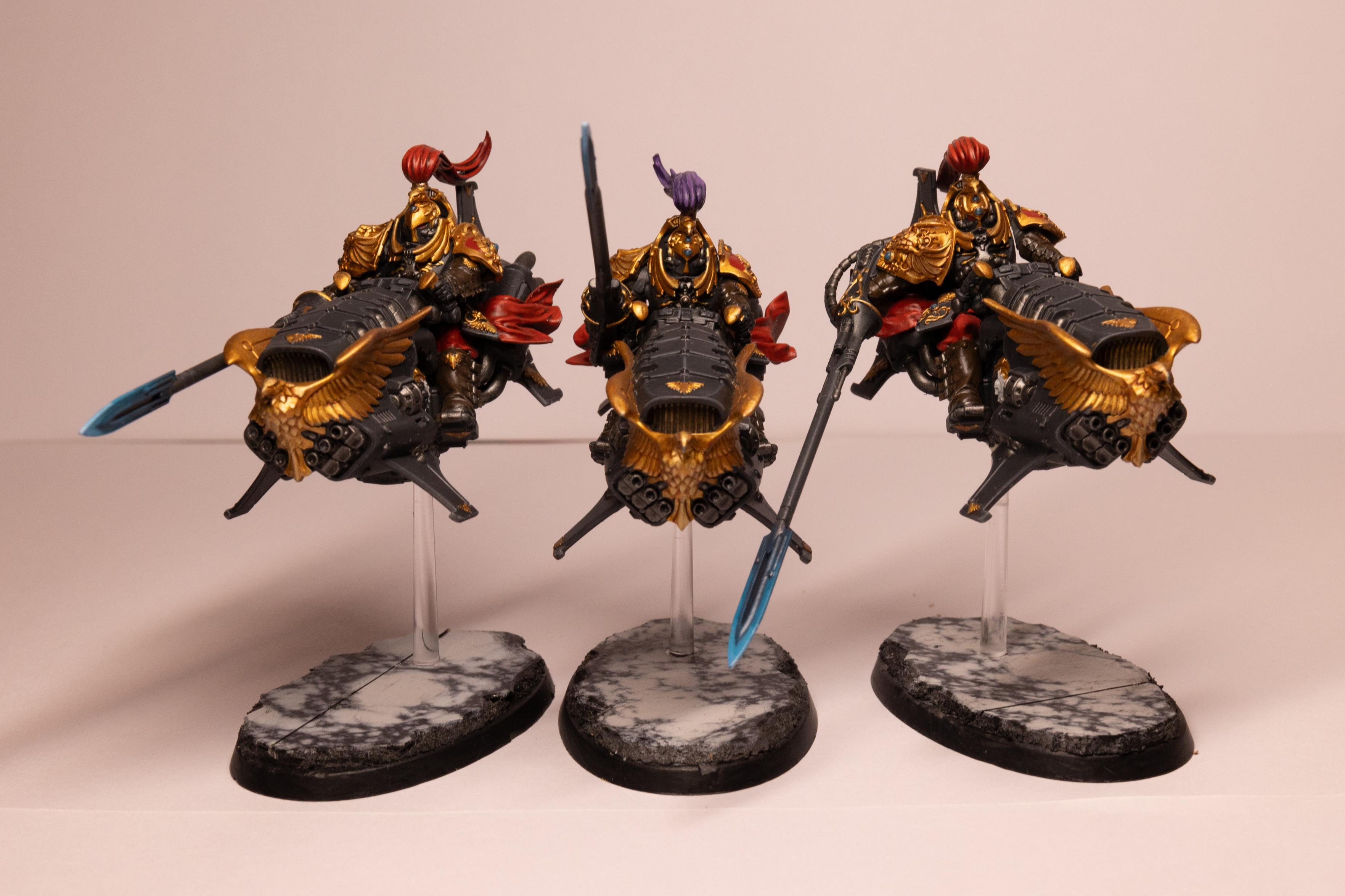 Three Warhammer 40k Custodian Vertus Praetors in the Shadowkeepers color scheme. They are three armoured warriors on large jet bikes painted in black armour with gold gold trim and red accents. The front of each bike features a large golden eagle and each warrior is holding a long lance with a glowing electric blue tip. The central warrior has a purple head plume. They are all mounted on clear plastic sticks on top of a black and while marble-like base